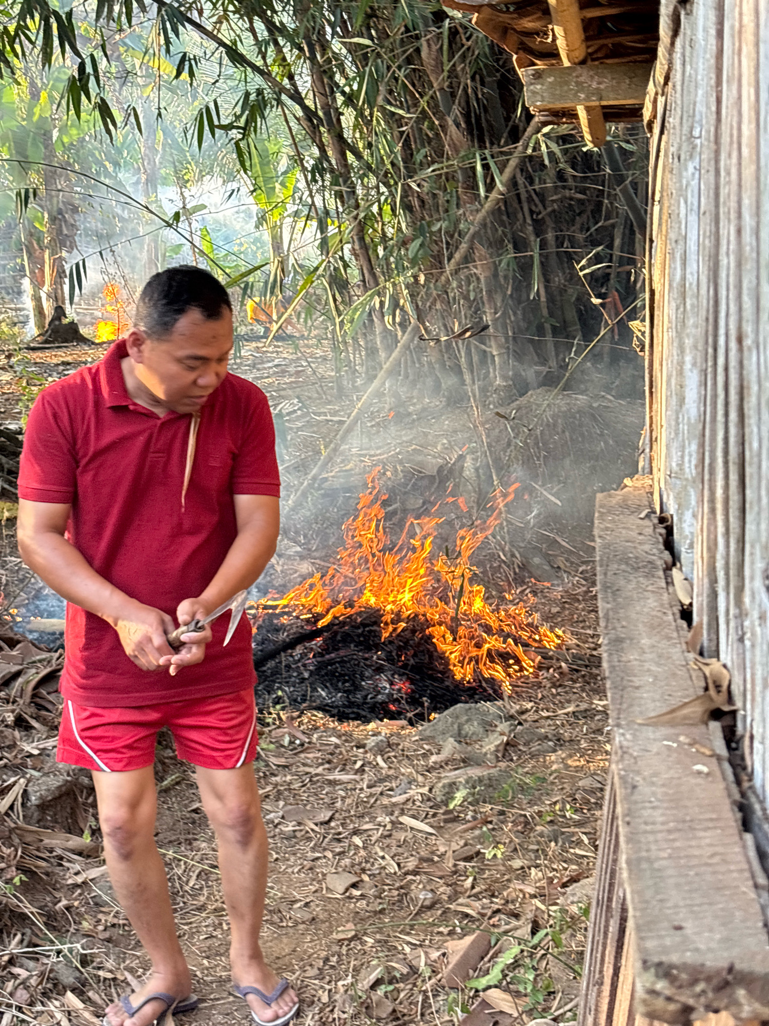 A man wearing a red polo shirt and red shorts stands in front of a small bonfire in an outdoor setting, possibly in Tirtomoyo, Indonesia. He is holding a tool with a blade in his hands. The fire is burning on dry leaves and debris on the ground, with smoke rising into the air. Behind the man and the fire, there are dense bamboo trees and other vegetation. To the right of the image, a weathered wooden structure is visible. The ground is covered in dry leaves, twigs, and some stones. The lighting suggests it is either early morning or late afternoon, with sunlight filtering through the foliage.