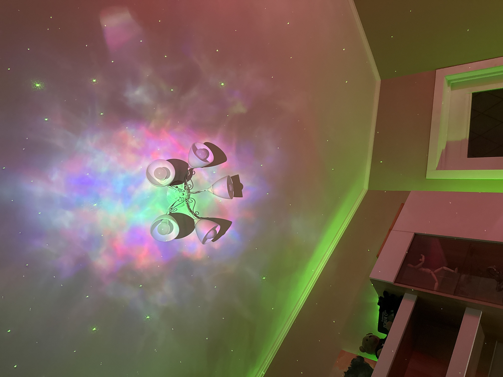 The image presents an upward view within an indoor room, showcasing a vibrant celestial projection on the ceiling and upper walls. The ceiling is awash with swirling, nebula-like patterns in hues of pink, purple, blue, and green, dotted with numerous small, bright green star-like points.
A white metal chandelier, featuring five light-colored, cup-shaped lamps, hangs prominently from the center of the ceiling, partially illuminated by the green light. The right side of the image reveals a light-colored wall, featuring a white-framed window with a dark pane, strongly outlined by a brilliant green light. Below the window, a portion of a white shelving unit is visible. One shelf displays an articulated, mannequin-like figure, while another lower shelf holds a small teddy bear and a black item with the partially visible text "POKER C".
The overall scene suggests a darkened room, likely at night, where a projector is creating an immersive, dreamy atmosphere. No people are visible.