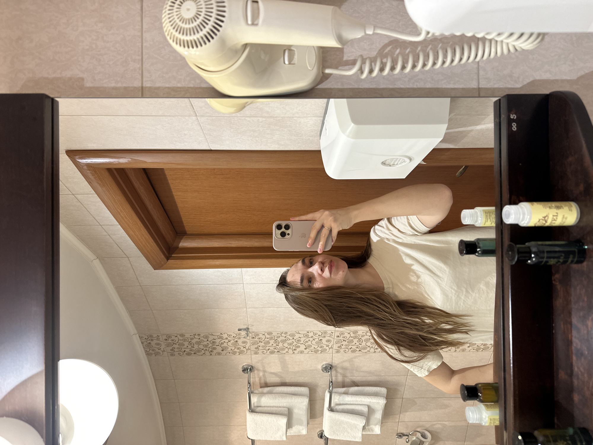 An indoor mirror selfie captures a young woman in what appears to be a hotel bathroom. The image is oriented with the top of the mirror to the left and the bottom to the right, showing the scene rotated 90 degrees clockwise.
The central subject is a woman with long, straight brown hair, wearing a light-colored t-shirt. She is holding a smartphone with a light-colored case, featuring a visible Apple logo and triple camera setup, to take her reflection's photo. Her face is directed towards the phone.
The bathroom setting is evident from the tiled walls, a white toilet bowl partially visible in the lower left of the reflection, and mounted fixtures. Reflected in the mirror are two towel racks with neatly folded white towels below patterned accent tiles. A wooden door frame is prominent in the reflection, and a white hand dryer is mounted on the wall above it. Above the mirror itself, a white wall-mounted hairdryer with a coiled cord is visible.
To the right of the mirror, a dark wooden shelving unit or cabinet holds several small amenity bottles, typical of hotel provisions. Some bottles are dark green/black, while others are clear or yellowish. Visible text on one bottle includes "TEL", likely part of "HOTEL". On the dark wooden shelf, the characters "∞ S" are visible. The lighting suggests daytime indoors or well-lit artificial lighting.