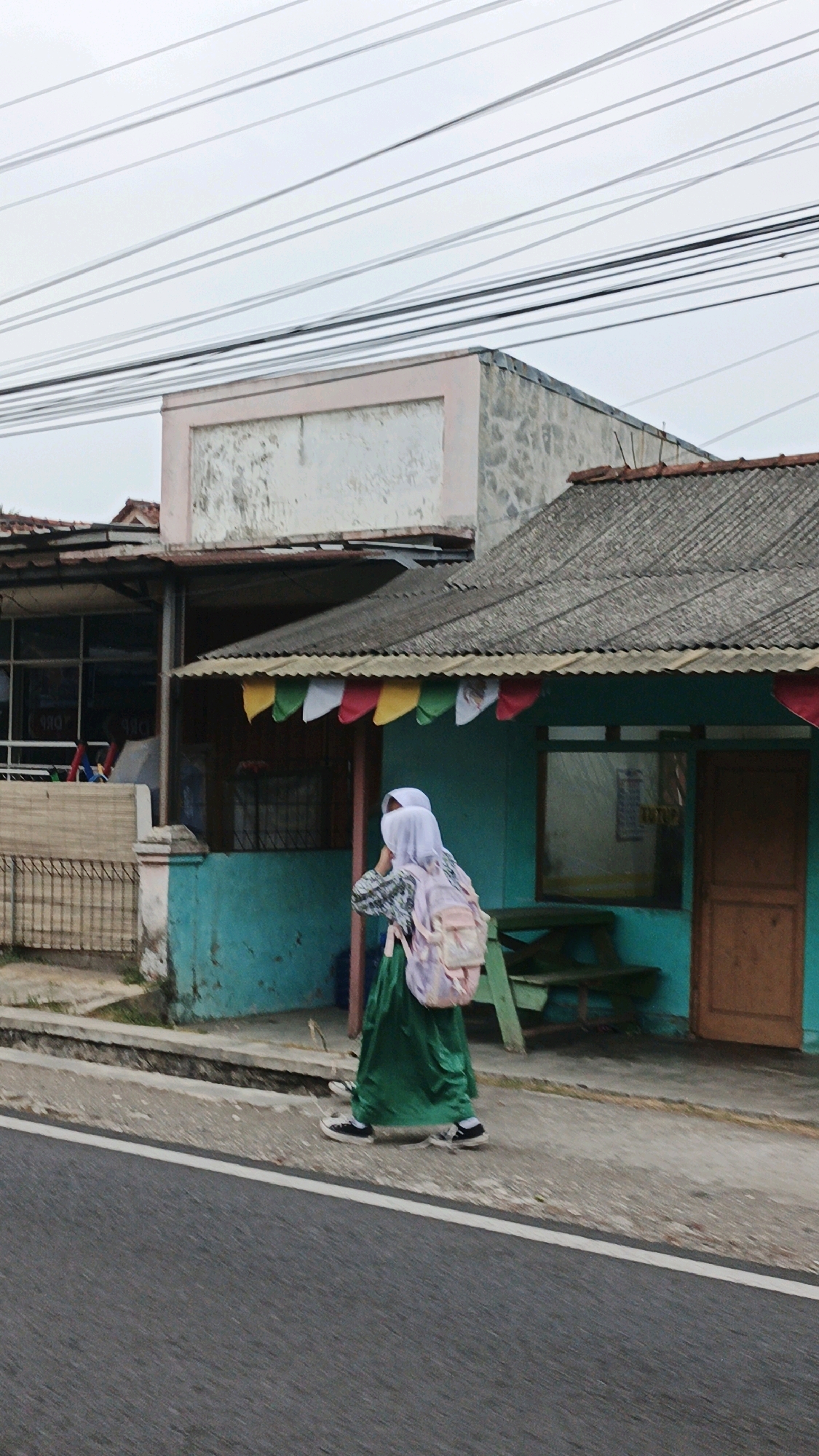 A person wearing a white hijab and a green skirt with a pink backpack walks on the side of a road in front of a turquoise building with a corrugated roof in Sukaresik, Indonesia. The building has a wooden door and a window. Colorful flags are strung along the edge of the roof. The sky is overcast.