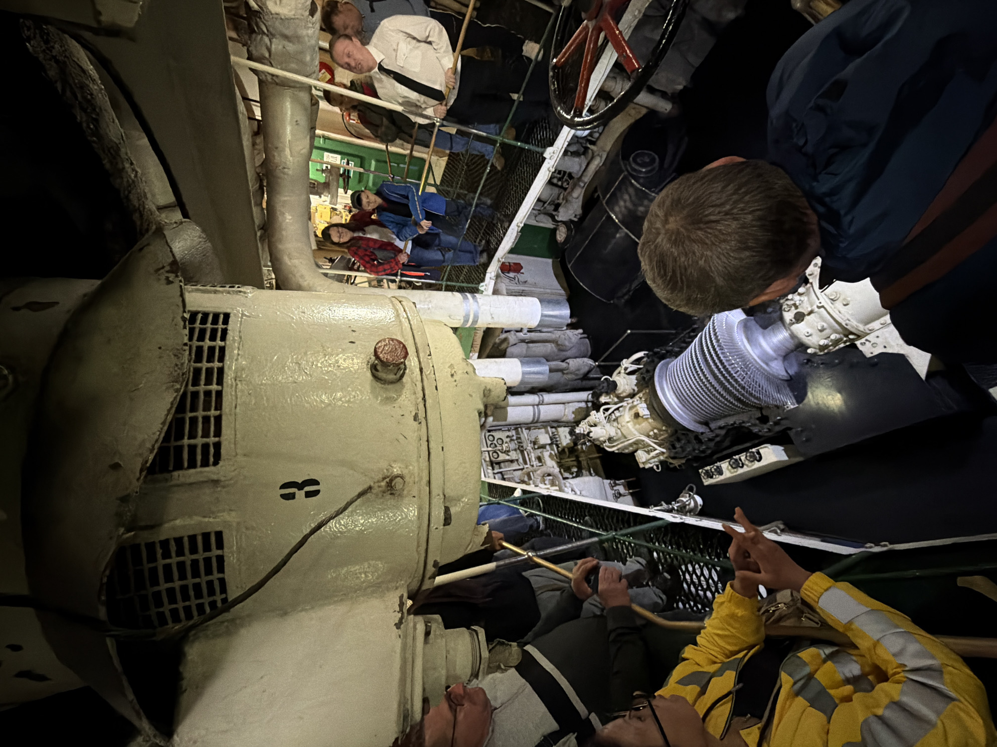 The image captures an interior view of the engine room of a vessel, likely docked in Murmansk, Russia. A group of people, including men, women, and children, are observing a large piece of machinery. The machinery appears to be an engine or a similar industrial apparatus, with prominent ribbed components and complex piping. The environment is utilitarian and industrial, with exposed pipes, metal grating floors, and visible structural elements of the ship. Some individuals are dressed casually, while one man in the foreground wears a formal shirt and tie, possibly acting as a guide or presenter. The lighting is somewhat dim, typical of an engine room, with artificial light sources highlighting the machinery and the people. The presence of a steering wheel on the right side of the frame further confirms the maritime setting. The overall impression is of a guided tour or an educational visit within a historical or operational ship.