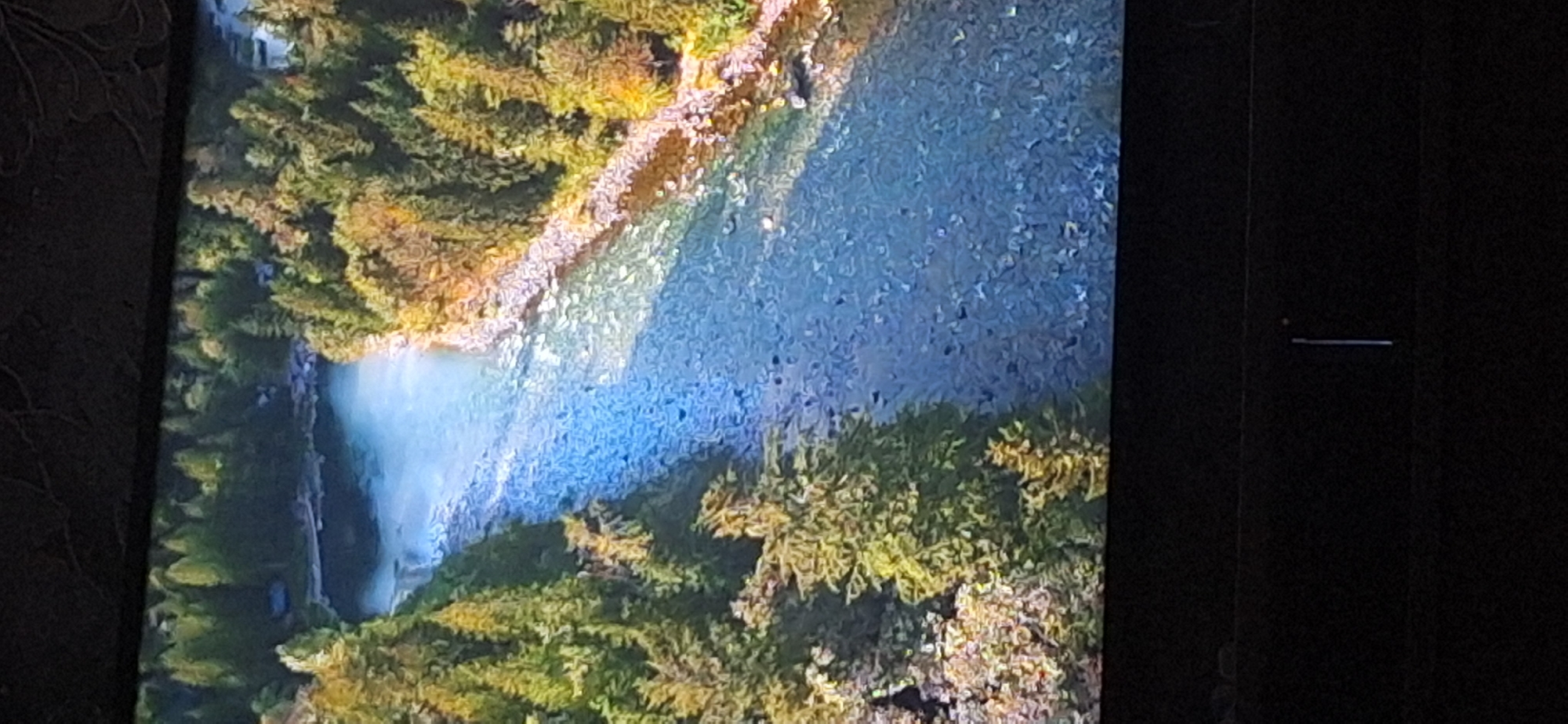 The image displays a wide, shallow river flowing through a forested area. The riverbed is rocky and visible through the clear, blue water. Dense green trees line both banks of the river, with some appearing to be pine trees. The overall scene suggests a natural, undisturbed landscape. There is no indication of the city of Bila Tserkva, Ukraine, within the image.