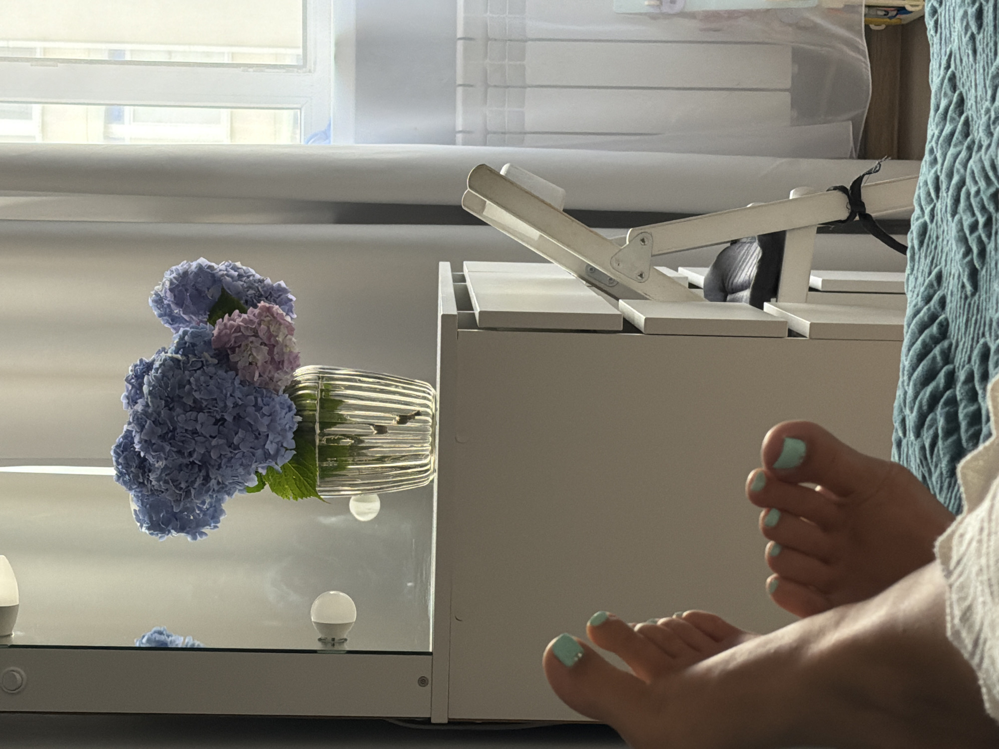 This media file features a close-up view of a person's feet, adorned with light blue nail polish. The feet are positioned on a light-colored surface, and another person's feet, also with light blue nail polish, are visible in the foreground, suggesting a shared relaxed moment.
In the background, a bouquet of blue and pink hydrangeas rests in a ribbed glass vase on a reflective surface, likely a table or shelf. This surface is part of a light-colored, minimalist furniture piece. A white adjustable stand is also visible, angled upwards. The scene appears to be indoors, near a window that is letting in a bright, diffused light, possibly indicating daytime, and a comfortable indoor setting. The overall mood is serene and tranquil.
There is no visible text in the media file. The location context is Sochi, Russia.