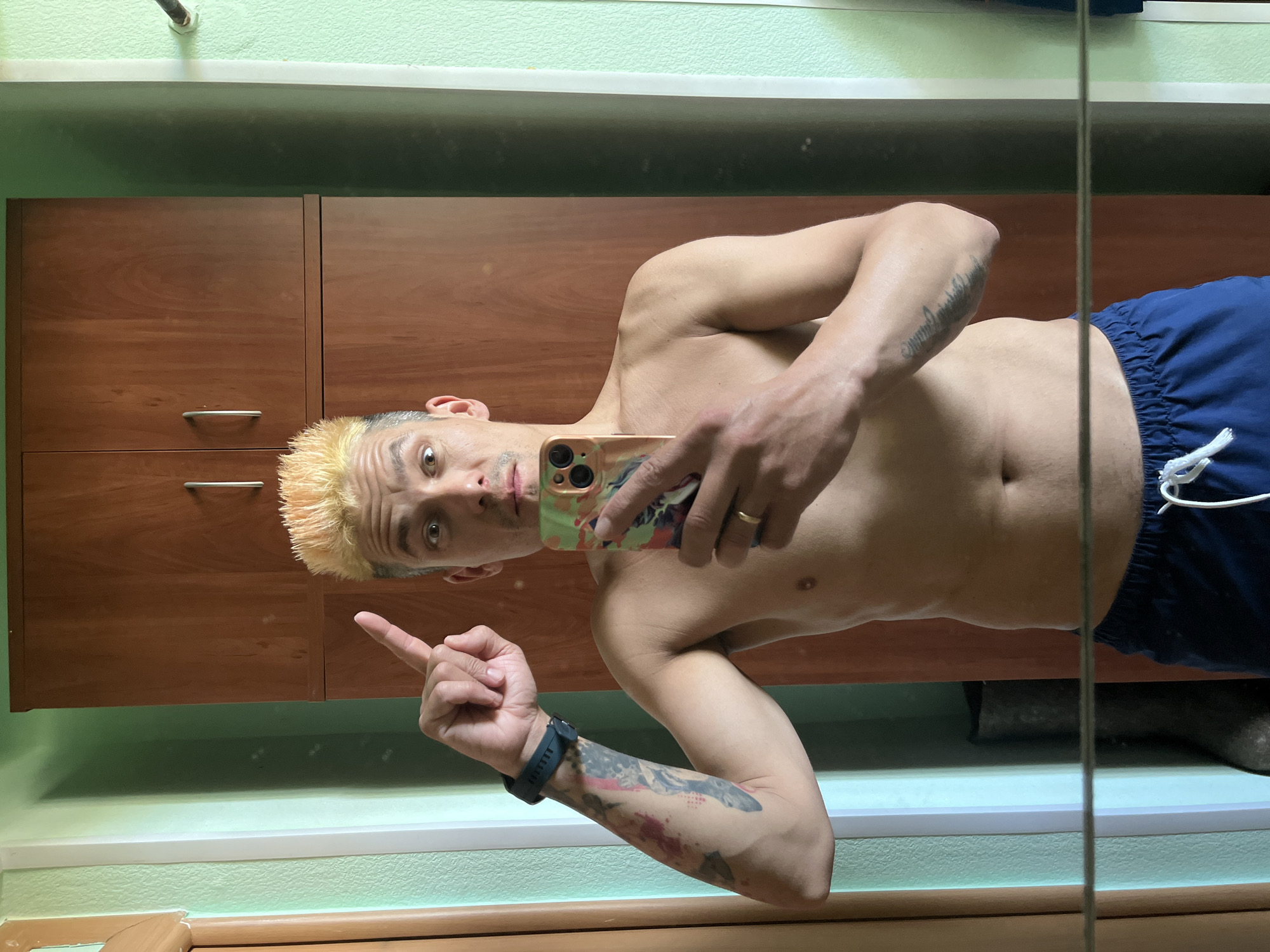 A man with bleached blonde hair, facing the camera, is taking a selfie in a bathroom in Chaikovsky, Russia. He is shirtless and wearing blue shorts. He is pointing with his left hand and holding a mobile phone with a colorful case in his right hand. The reflection shows a cabinet behind him, a green wall, and wooden trim. A small piece of a vertical support is visible near the right edge of the image, likely part of a mirror frame. A pipe is also visible on the ceiling near the top of the frame. The man has tattoos on his arms and is wearing a dark-colored fitness tracker on his left wrist.