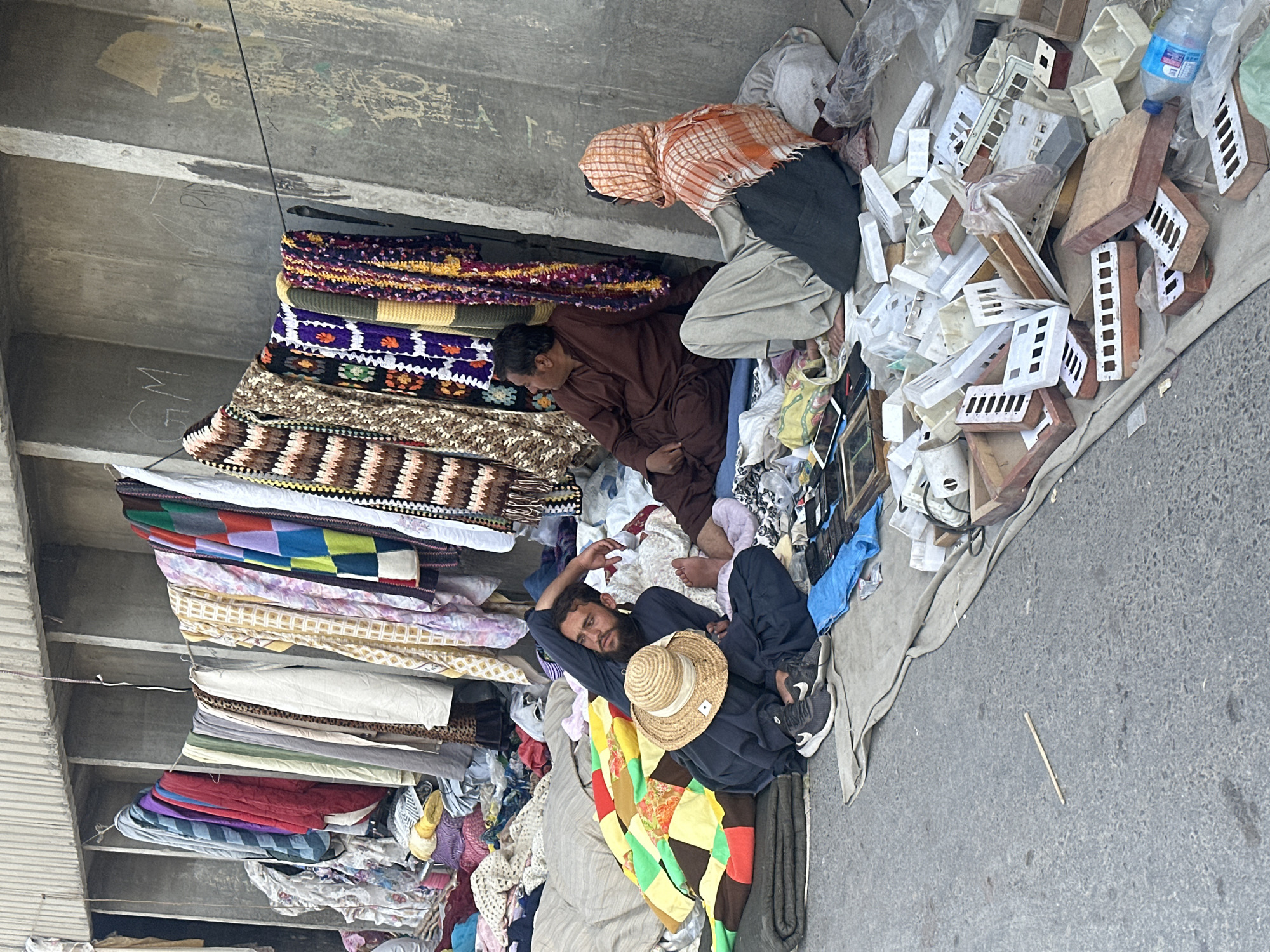 In Quetta City Tehsil, Pakistan, a street vendor lies amidst stacks of textiles and other goods, apparently resting. He wears a dark tunic, pants, sneakers, and a straw hat. Next to him is a stack of colorful patchwork quilts. Behind him, a variety of stacked textiles, including blankets and fabrics, rise toward an overhead concrete structure. Another man sits behind the textiles, wearing brown clothing. To the right of the resting man, another vendor kneels sorting through various white and tan plastic electrical boxes. He wears a light-colored turban and dark pants. Trash and scattered items are seen on the ground. The scene is a street or alleyway.