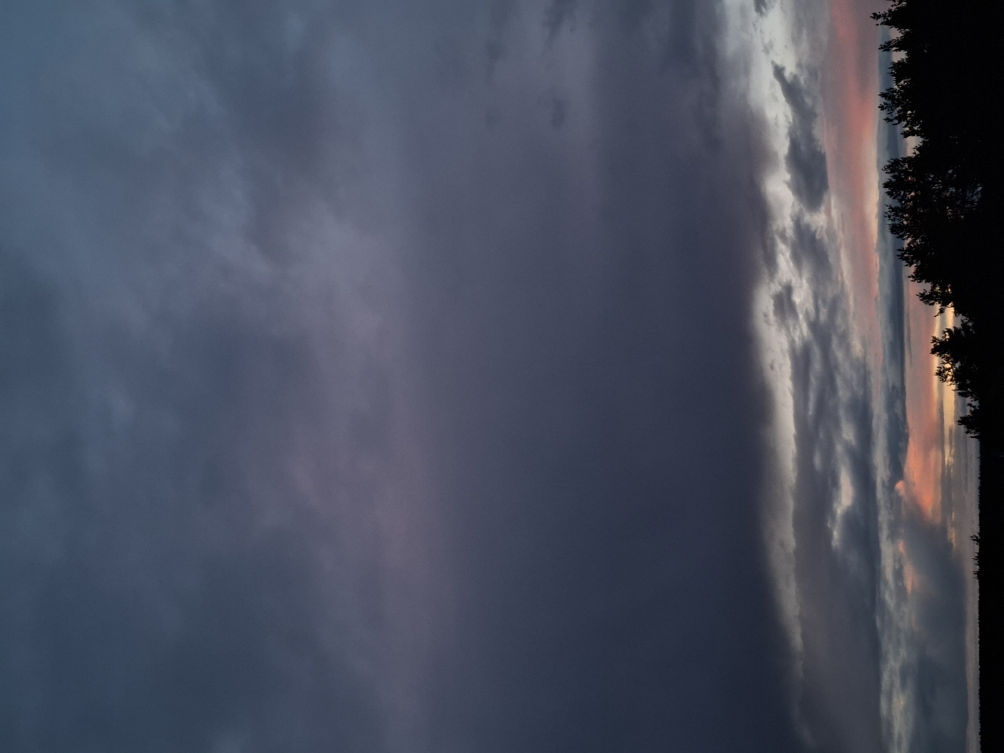 The image shows a sky at dusk. Dark, heavy clouds fill the left side of the frame. Towards the right, lighter clouds transition into shades of pink and orange, indicating the sunset or sunrise. The silhouette of trees appears along the top right edge, suggesting the presence of a forest or wooded area. The image's location is Khlevnoye, Russia, but no other specifics within the town or the presence of people, subjects, objects, actions, or interactions are evident.