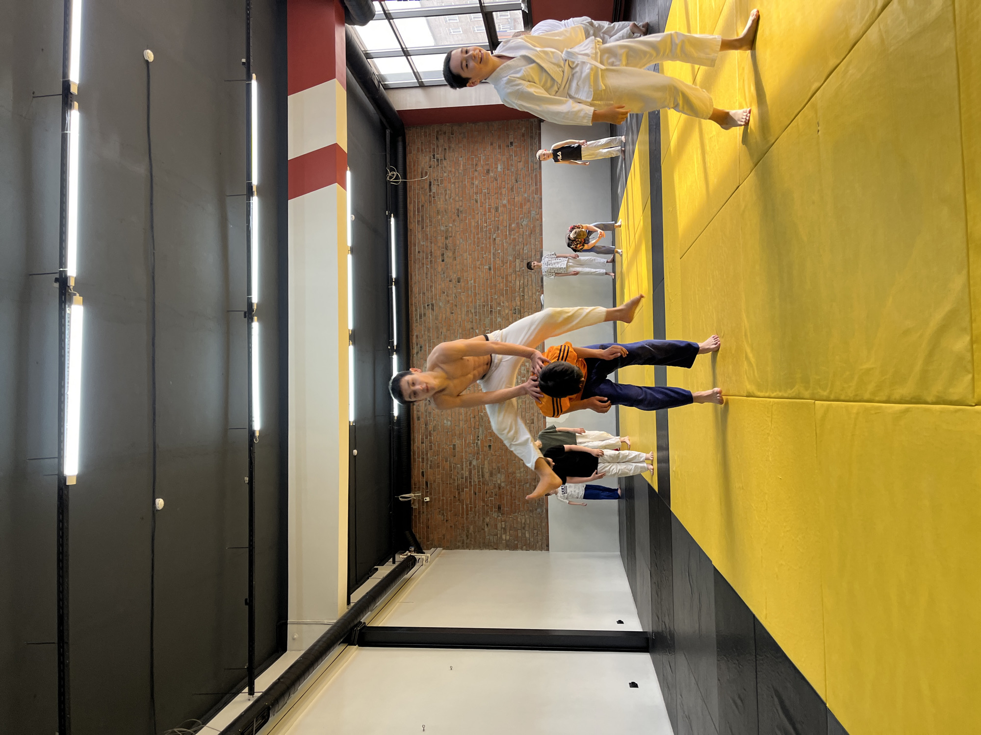 The image shows an indoor martial arts training area. Several people are present, some in martial arts uniforms and others in casual clothing. They are on a padded mat floor that is yellow and black. The walls are a combination of black, brick, and yellow-striped surfaces. There are linear fluorescent lights on the ceiling and along the walls. Based on the location information, this scene is in Krasnoyarsk, Russia. The people appear to be engaged in a martial arts training session, possibly Judo. One person is lifting another.