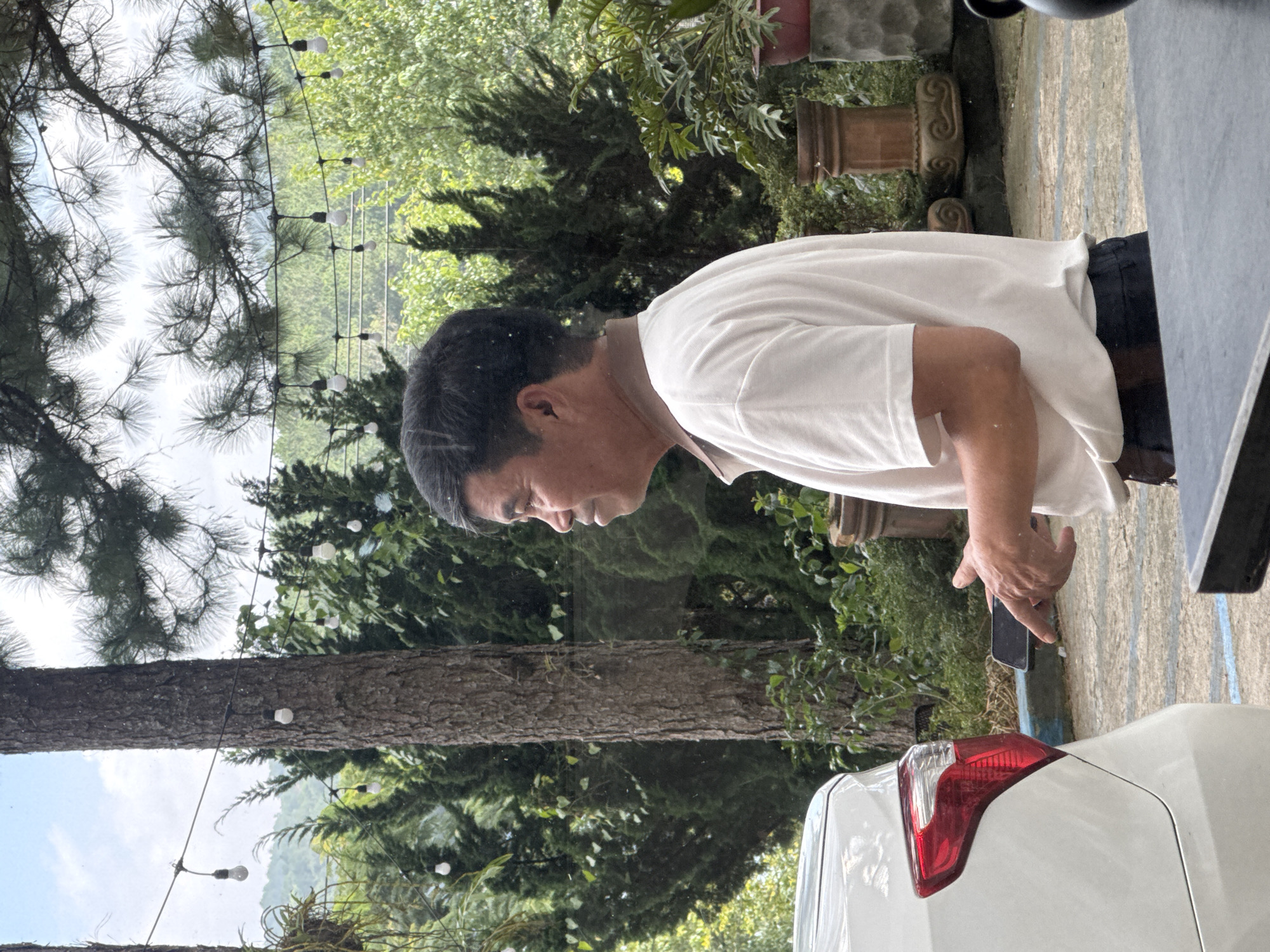 An East Asian man, appearing to be in his late 50s to early 60s, stands outdoors in Da Lat, Vietnam. He is wearing a white polo shirt and dark pants, and is holding a black smartphone in his right hand. The man is looking to his left, with his head slightly turned. In the background, lush green trees and foliage are visible, suggesting a natural or garden setting. String lights are hung between trees, indicating a decorative or functional lighting setup. A portion of a white car is visible on the left side of the frame, and decorative planters can be seen in the mid-ground. The ground appears to be paved with stone tiles. The overall scene depicts a man in a relaxed, outdoor environment, possibly a cafe, resort, or residential area in Da Lat known for its temperate climate and abundant greenery.