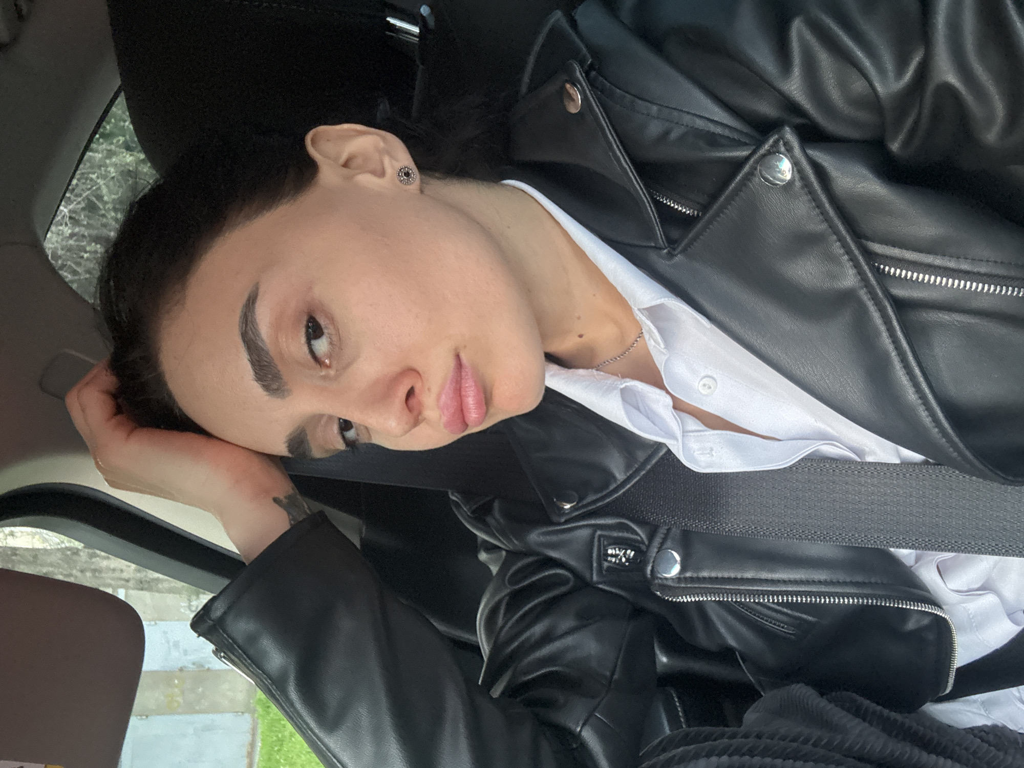 A woman is in a car wearing a black leather jacket over a white button-down shirt, with a black seatbelt across her chest. She is resting her head on her hand. The car appears to be parked; the background shows glimpses of trees and buildings through the window. This cannot be used to locate the exact location within Tbilisi.
