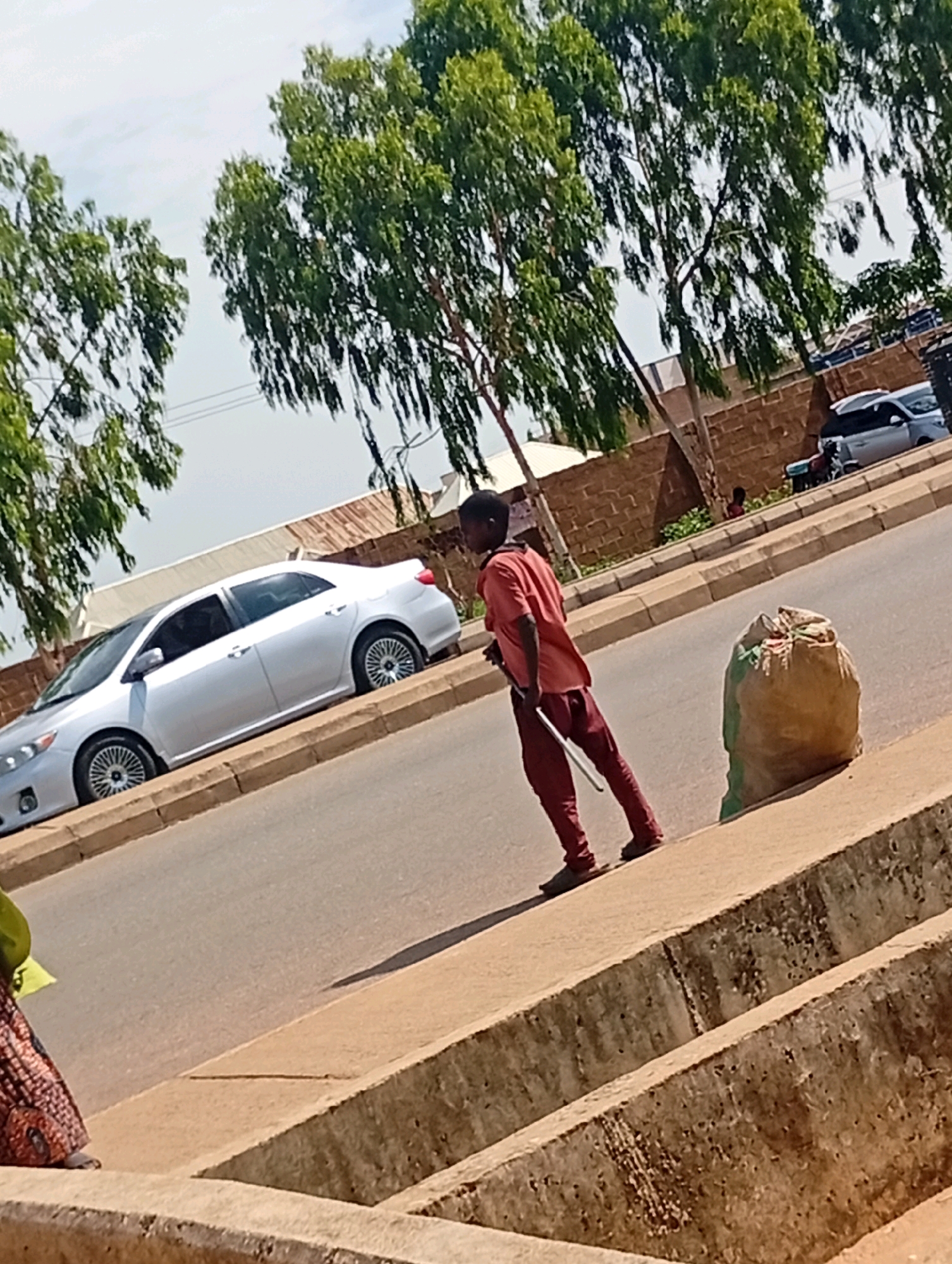 In Tumfure, Nigeria, a young male stands on a paved road, holding a long, thin object that appears to be a stick or broom handle. He is dressed in a red t-shirt and dark red trousers. Next to him on the sidewalk is a large, roughly spherical bag, possibly filled with trash or produce. In the background, a silver sedan is parked on the side of the road, and beyond that, a brick wall and trees line the street. The sky is visible and appears cloudy. The scene depicts a typical urban setting with elements of daily life.