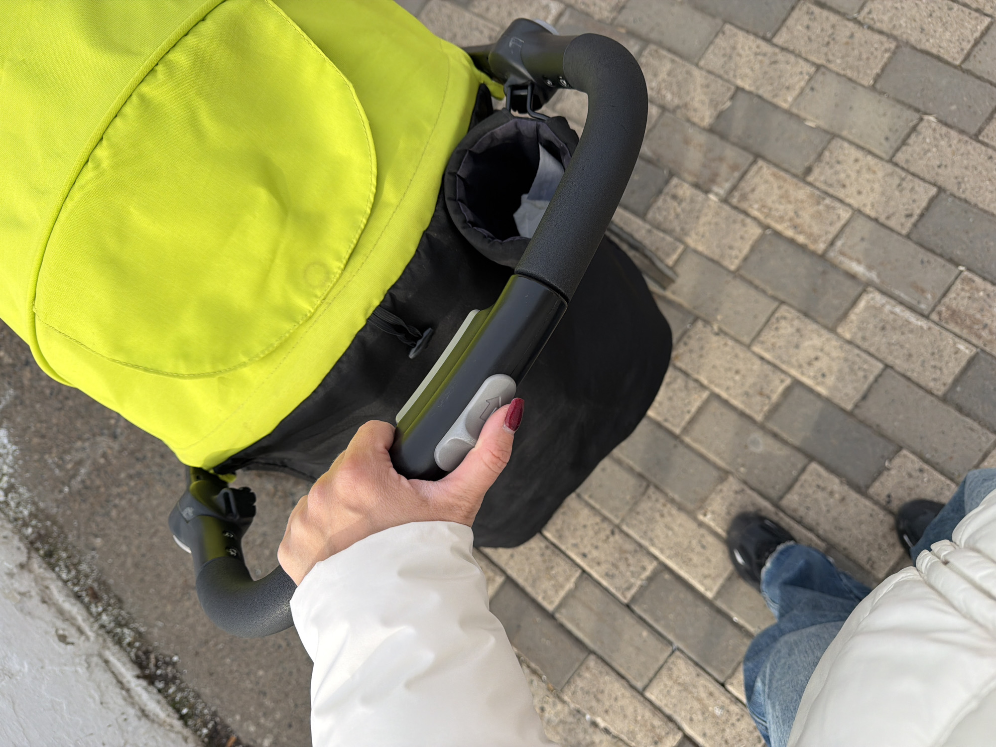 The image shows a person pushing a stroller on a paved walkway. The stroller has a bright yellow fabric top. The person is wearing blue jeans, black shoes, and a white jacket. The location is Votkinsk, Russia, as the image was taken there.