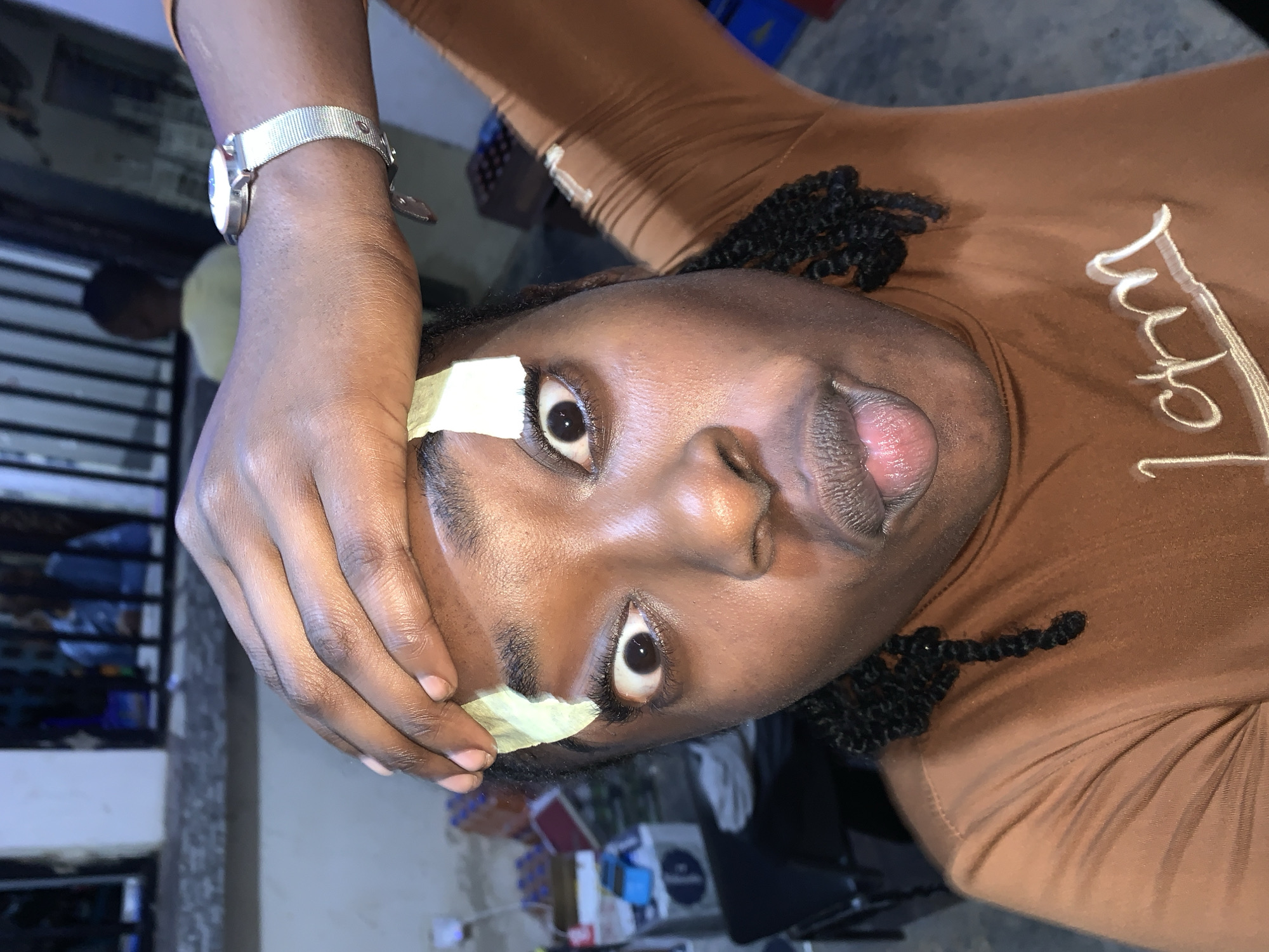 A medium-shot portrait captures a person with dark skin and braided hair in New Stadium, Nigeria. The individual wears a brown long-sleeved top with a high neck, featuring a white embroidered design on the chest that appears to read "Lctm". A silver-toned watch with a mesh band is visible on the right wrist. The person's right hand is raised and placed on their forehead, with fingers resting above the eyebrows. Two rectangular pieces of light green adhesive tape are affixed to the forehead, positioned above the inner part of each eyebrow. The subject's eyes are wide open with dilated pupils, directly engaging the camera. Their mouth is slightly open, with the tongue partially extended. The background suggests a semi-outdoor environment, featuring a dark metal gate or railing. Various objects, including plastic bottles and a blue container, are visible on the ground to the left. To the right, stacked items resembling boxes or crates are present. A blurry figure of another person in a light-colored shirt is discernible in the background, behind the gate, facing away from the camera.