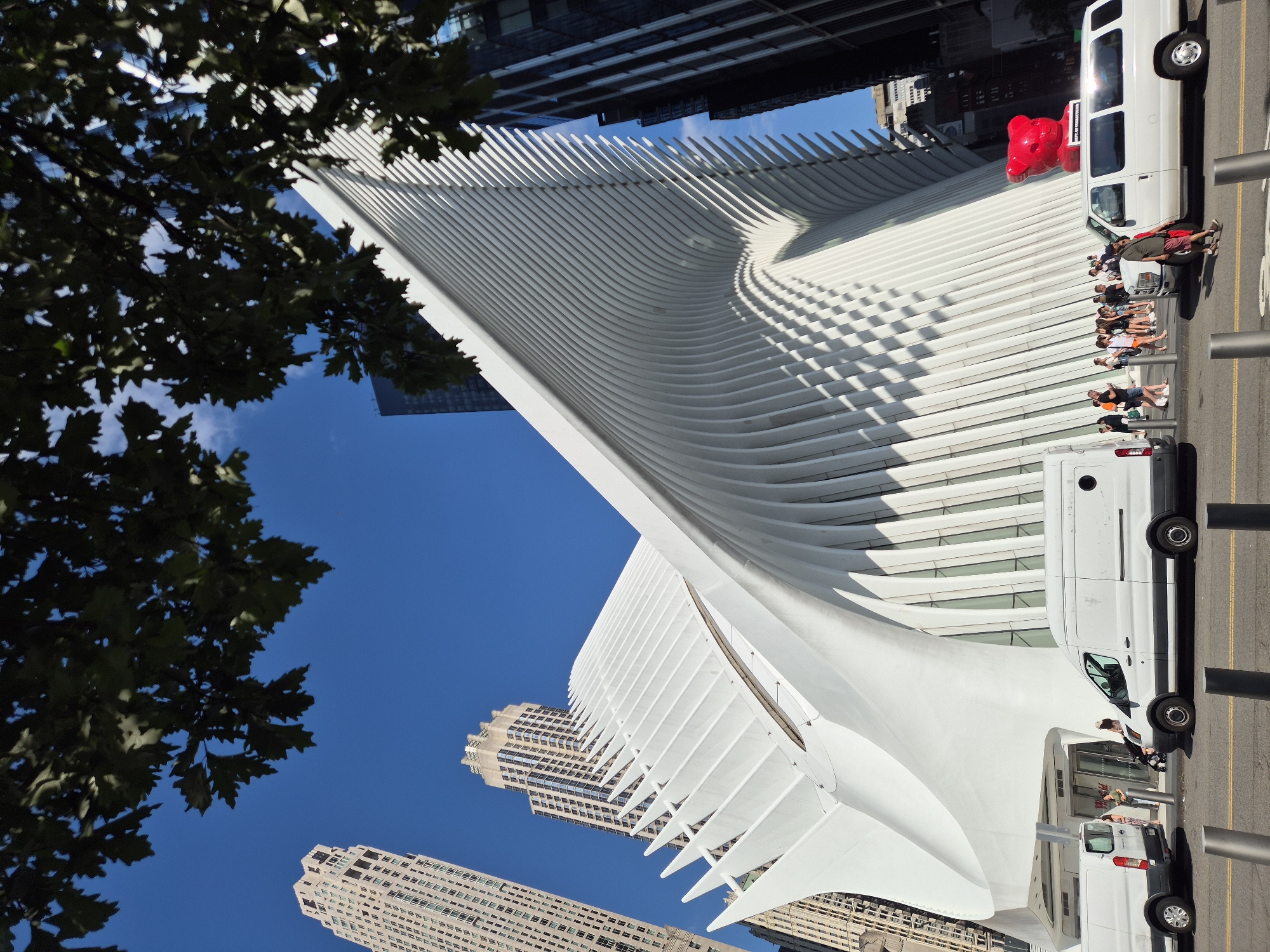 The image shows the Oculus transportation hub in New York City. The white, winged structure of the Oculus dominates the foreground, its ribbed roof casting dramatic shadows on its facade. To the left of the Oculus, two tall, beige skyscrapers are visible against a clear blue sky. In the midground, several white vans are parked on a street in front of the Oculus. People are seen walking and standing near the entrance of the building, and some are near the vans. A bright red, bear-shaped sculpture with text on its base is perched atop one of the vans. Trees with dark green leaves are visible in the upper portion of the image, partially obscuring the sky. The scene is lit by bright sunlight, suggesting it is daytime.