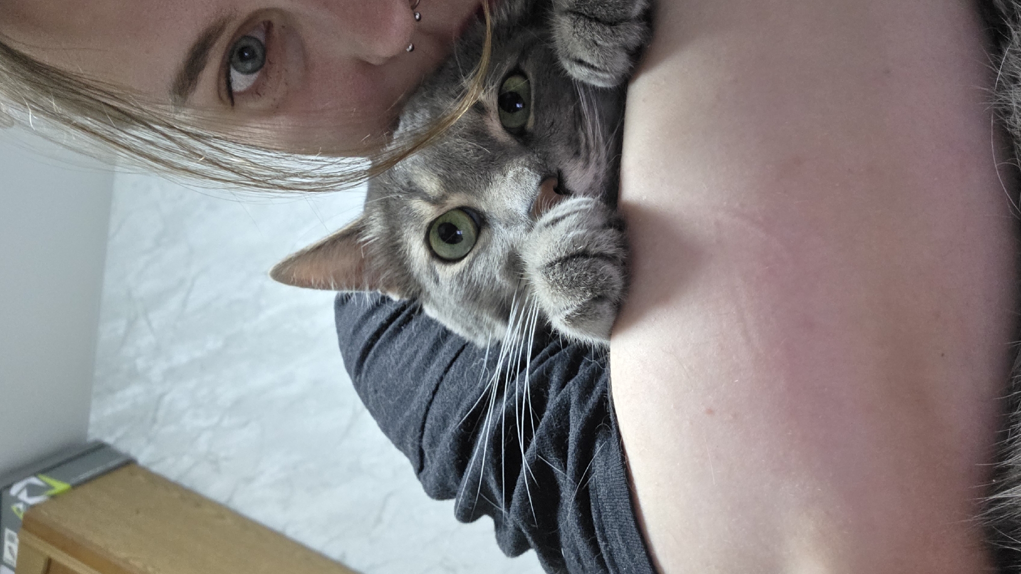 A close-up, slightly rotated portrait shot shows a person holding a gray tabby cat. The person's face is visible in the upper left corner, with light-colored eyes and blonde hair partially obscuring their forehead. They have a lip piercing. The cat is held close to the person's body and looks at the camera with green eyes. The cat's paws are near the person's face. The person wears a dark gray or black shirt. The background is a light-colored wall with a cabinet in the lower left corner. The location is Unknown, Unknown.