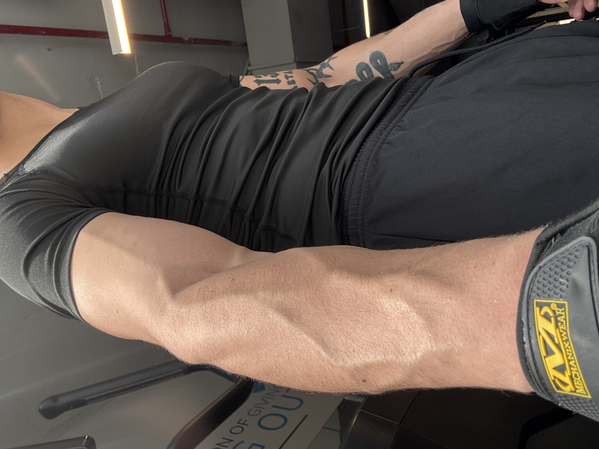 The image depicts a muscular arm with prominent veins, clad in a black compression shirt and black shorts. A portion of a workout glove labeled "Mechanix Wear" is visible on the wrist. Tattoos adorn the forearm. The background suggests a gym setting, with a piece of exercise equipment visible and text on the equipment that reads "...ON OF GIVING...OU". Given the limited contextual clues, it's impossible to determine the location in Sofia, Bulgaria.