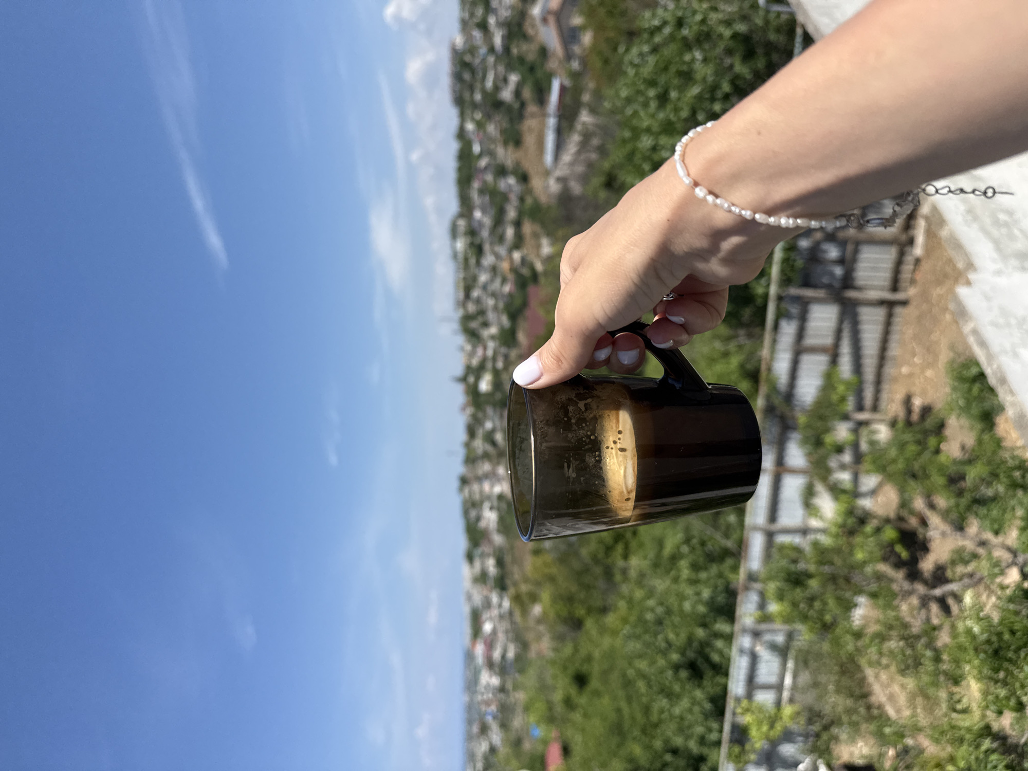 A close-up view from an elevated perspective captures a right human hand holding a dark, semi-transparent glass mug filled with coffee, topped with a light brown crema. The hand, adorned with white-tipped fingernails, wears a bracelet composed of small white pearls, with a silver chain and charm hanging below the wrist. The cup is positioned against a background featuring a sprawling, distant city or townscape with numerous buildings and abundant green trees and foliage, indicative of a sunny outdoor environment. In the mid-ground, below the cup, a concrete ledge is visible, followed by a metal fence line partially obscured by dense greenery. The upper portion of the frame is dominated by a clear, bright blue sky with sparse, wispy white clouds. The scene is located in the city of Unknown, Unknown.