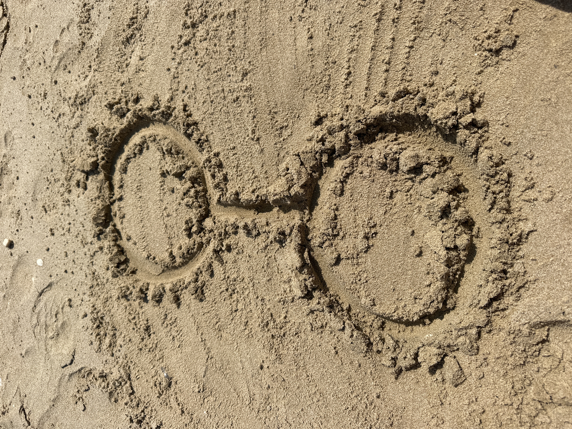 The image shows a drawing in the sand, resembling the symbol for Venus. The drawing consists of two circles connected by a short vertical line. The circles are not perfectly round, and the lines are somewhat uneven, indicating they were drawn by hand in the sand. The sand is light brown and appears to be wet in some areas, with some small pebbles and shells visible. The drawing is located on a beach in Pineto, Italy.