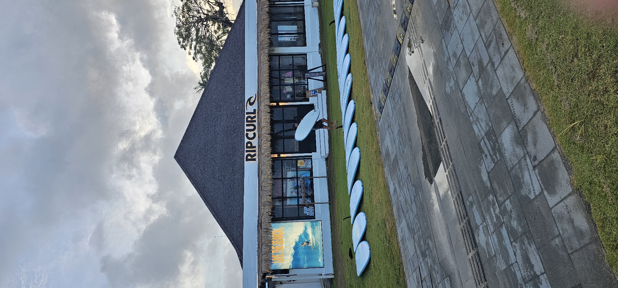 The image depicts a Rip Curl store in Denpasar, Indonesia. The store has a thatched roof and is adjacent to a row of surfboards displayed on the grass. A person is visible carrying a surfboard outside the store. Next to the grass, there is a gray tile pavement that shows signs of recent rain. The sky is overcast with clouds.