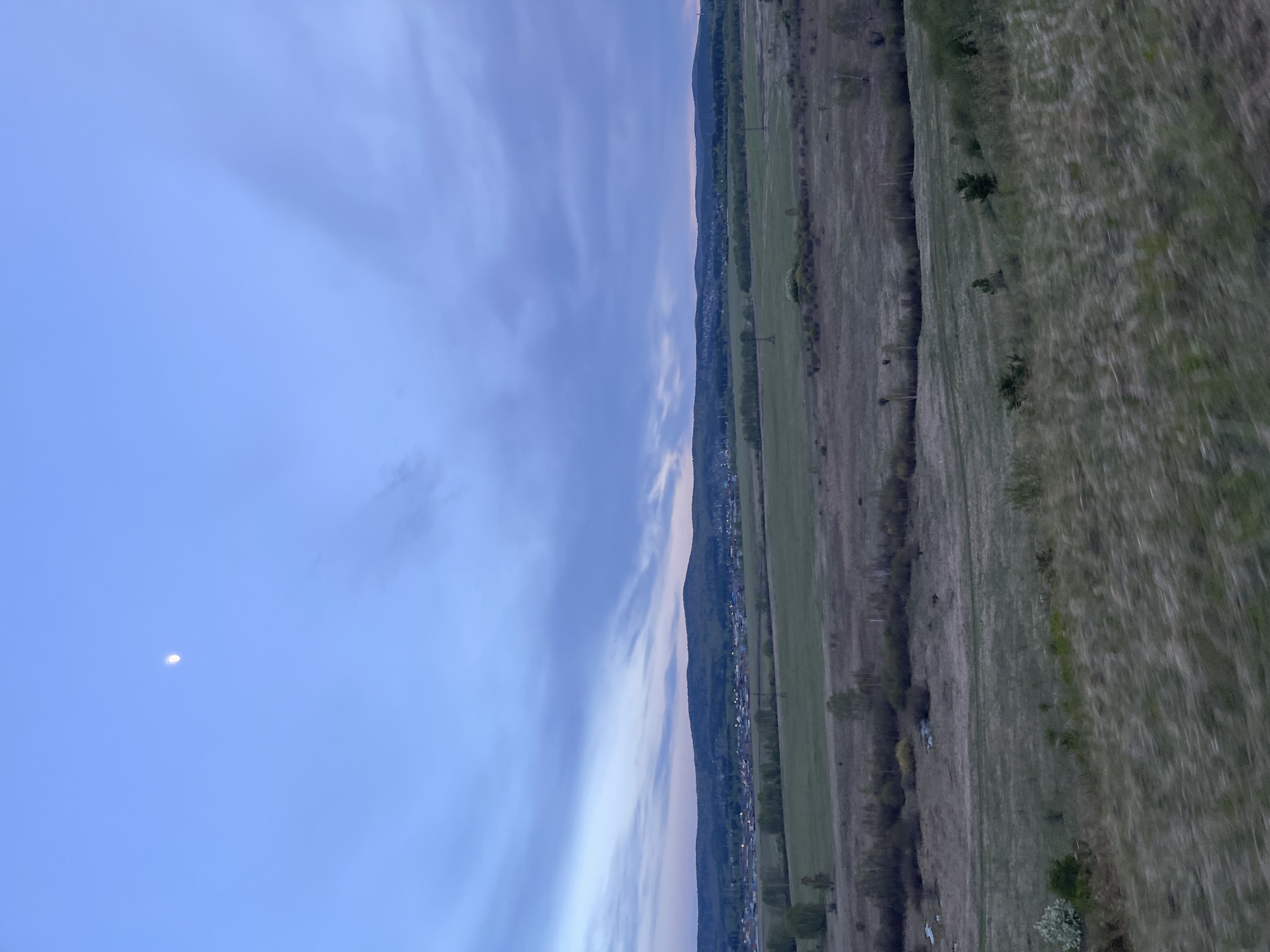 The image presents a landscape in the vicinity of Элита, Russia, under a twilight sky. A crescent moon is visible in the upper left. The scene is split between the sky and land. The land is a mix of fields and vegetation bordering the edge of the town of Элита. The horizon line is roughly in the middle of the image.