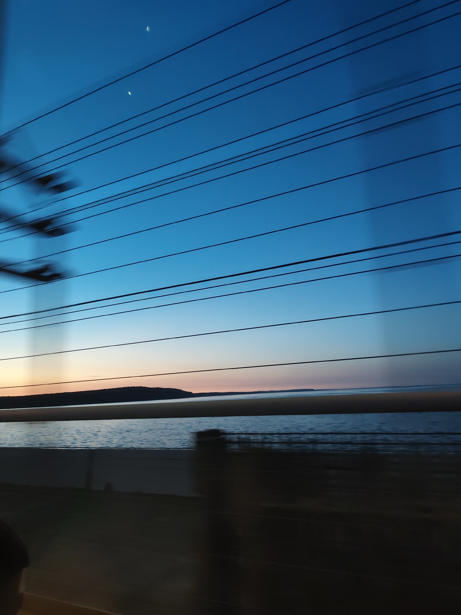 The image captures a scene viewed through a window, likely from a moving vehicle. The sky transitions from a deep blue at the top to a lighter hue near the horizon, with a hint of sunset colors. Power lines stretch across the upper portion of the frame. A body of water, possibly a lake or river, is visible in the middle ground, with land visible in the distance. The foreground is obscured by the interior of the vehicle and reflections on the window. The image appears to have been taken in a region with lakes and rolling hills. While I cannot definitively confirm the location as Chaikovsky, Russia without additional information or clues in the image, the landscape is consistent with the general geographic features of the region.