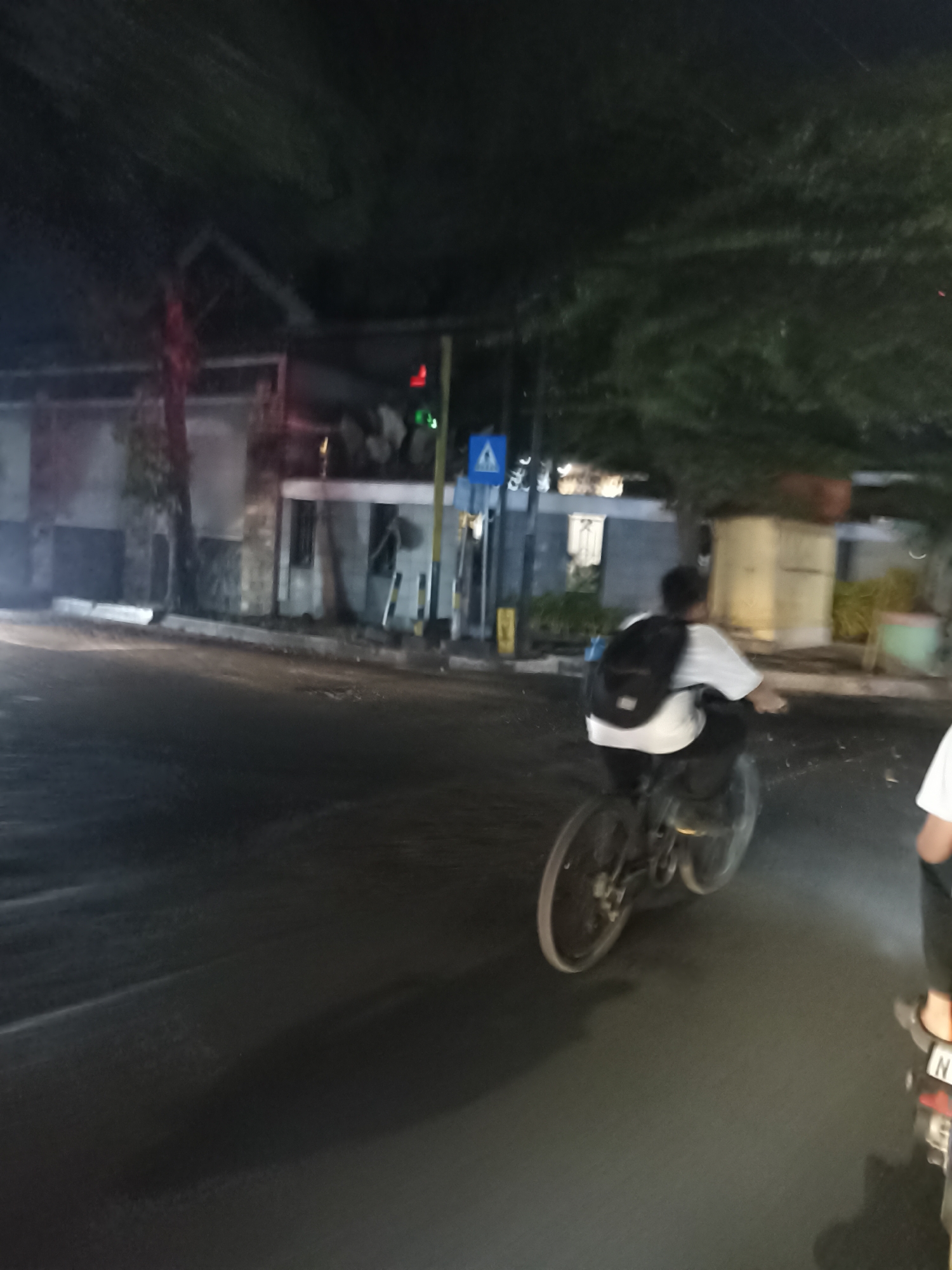 In Tompokersari, Indonesia, at night, a person wearing a white shirt and a black backpack rides a bicycle on a paved road. The road appears to be wet or reflective due to the lighting. In the background, there are buildings with some trees and foliage overhead. A traffic light is visible, as is a blue pedestrian crossing sign. Another person on a motorcycle is partially visible on the right side of the frame, with a license plate displaying "N". The lighting suggests an urban environment.
