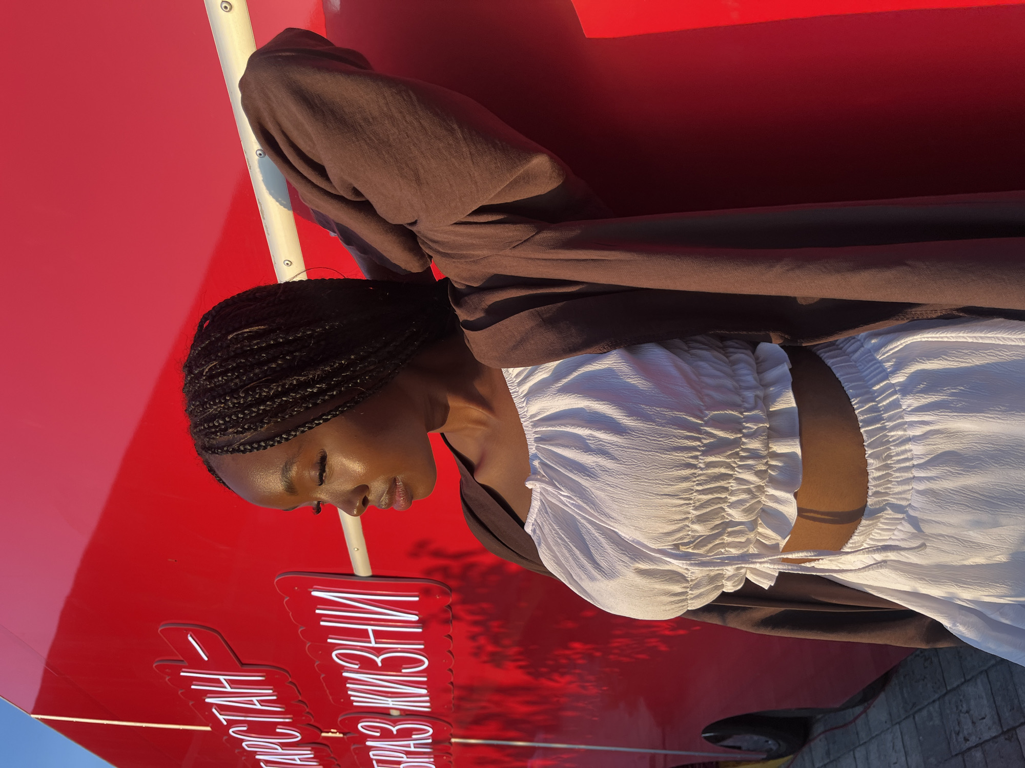 A young Black woman with braided hair is leaning against a bright red, curved surface. She is wearing a white ruffled crop top and white pants, with a brown open cardigan draped over her shoulders. Her left arm is bent with her hand resting on the back of her head. The red surface has white neon lettering that reads "ТАТАРСТАН" and "ОБРАЗ ЖИЗНИ" in Russian. The scene is illuminated by the warm light of the setting sun, casting a shadow on the ground. The backdrop appears to be a structure or vehicle located in Kazan, Russia, as indicated by the signage.