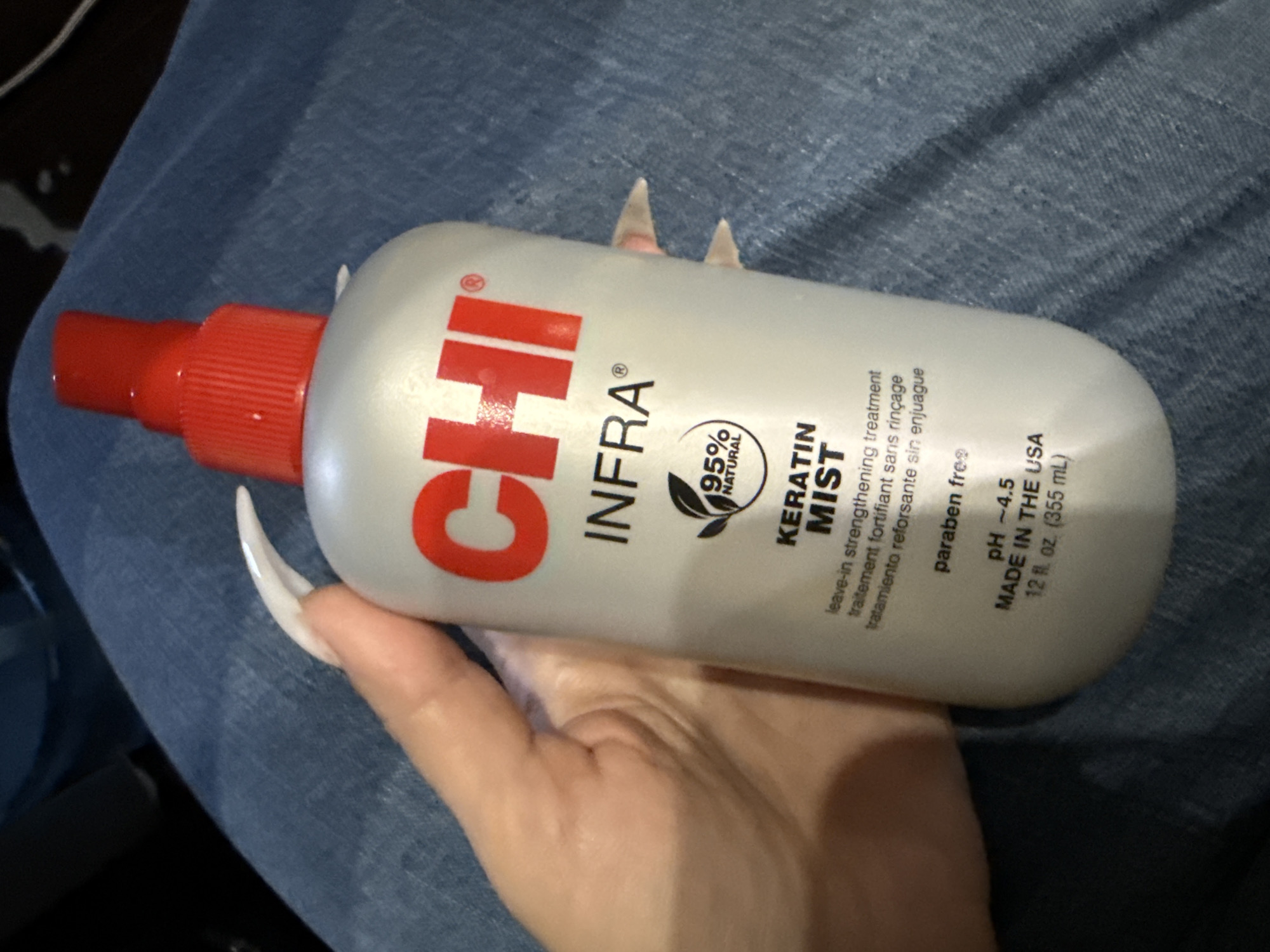 This media file features a close-up shot of a person's hand holding a bottle of CHI Infra Keratin Mist. The hand has long, white, pointed artificial nails, and is positioned against a backdrop of blue denim fabric. The bottle is white with red and black lettering. The text on the bottle is visible and reads "CHI INFRA®", "95% NATURAL", "KERATIN MIST", "leave-in strengthening treatment", "traitement fortifiant sans rinçage", "tratamiento reforsante sin enjuague", "paraben fre", "PH - 4.5", "MADE IN THE USA", and "12 fl. oz. (355 mL)". The lighting suggests an indoor setting, possibly with artificial light. There are no obvious cues to suggest a specific time of day, weather, or the location being Odesa, Ukraine. The overall impression is one of a product detail shot, focusing on the item and the person's hand.