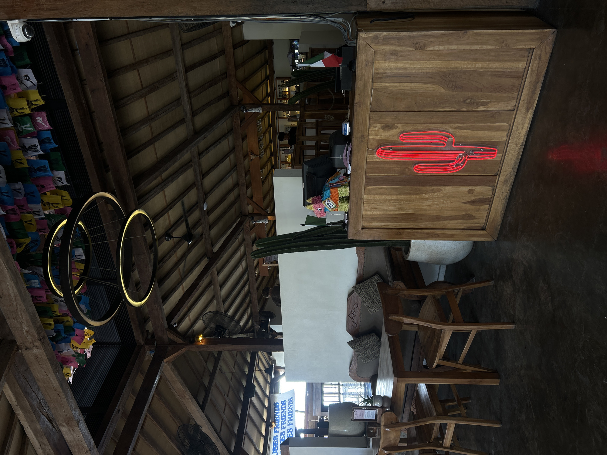 An indoor shot shows a restaurant in Kuta Selatan, Indonesia. In the foreground, a wooden counter has a red neon cactus sign and a small piñata. Behind the counter, a bartender is visible. To the left, a wooden table with chairs is set up. Above, exposed wooden beams support a thatched roof decorated with colorful flags. A modern ring-shaped chandelier hangs from the ceiling.