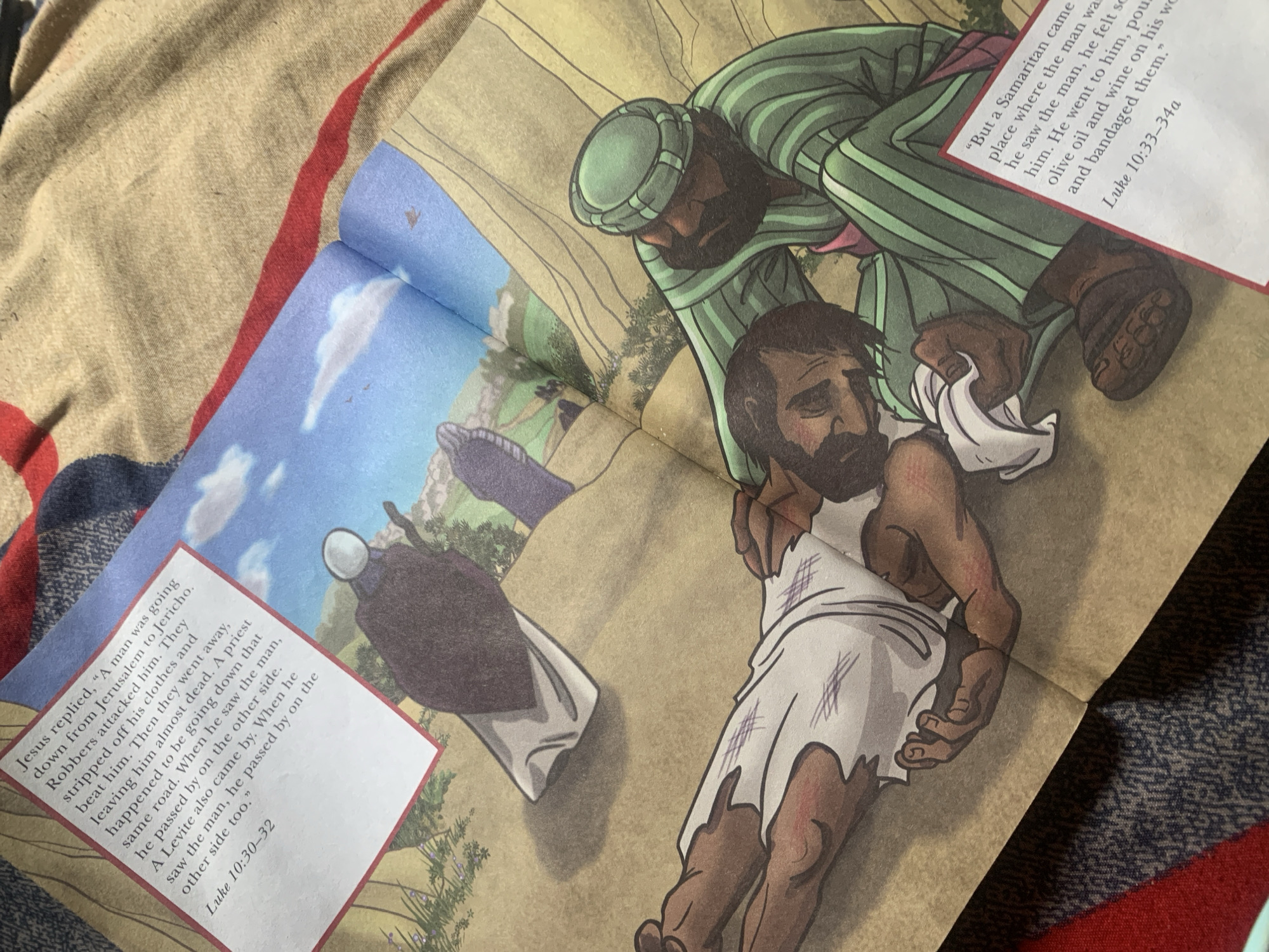 The image depicts an open book, likely a children's bible story, with illustrations and text. On the left page, text reads: "Jesus replied, “A man was going down from Jerusalem to Jericho. Robbers attacked him. They stripped off his clothes and beat him. Then they went away, leaving him almost dead. A priest happened to be going down that same road. When he saw the man, he passed by on the other side. When he saw the man, he passed by on the other side too." Below this text is "Luke 10:30-32."
The illustration on the left page shows a figure in dark robes walking away from a person lying on the ground, with barren hills in the background. The right page features an illustration of a wounded man lying on his side, dressed in torn white cloth. Another man, wearing a green striped robe and a green turban, is tending to him, holding a white cloth to his wounds. Text on the right page reads: "But a Samaritan came to the place where the man was. When he saw the man, he felt sorry for him. He went to him, poured olive oil and wine on his wounds and bandaged them." Below this is "Luke 10:33–34a."
The image is taken from an angled overhead perspective, showing the book lying on a textured, beige fabric surface, possibly a blanket or towel. Parts of a red and blue patterned fabric are visible at the edges of the image. The setting is not identifiable as Jauro Yinu, Nigeria, or any specific city. The scene is a biblical narrative illustration, not a depiction of a real-world location.