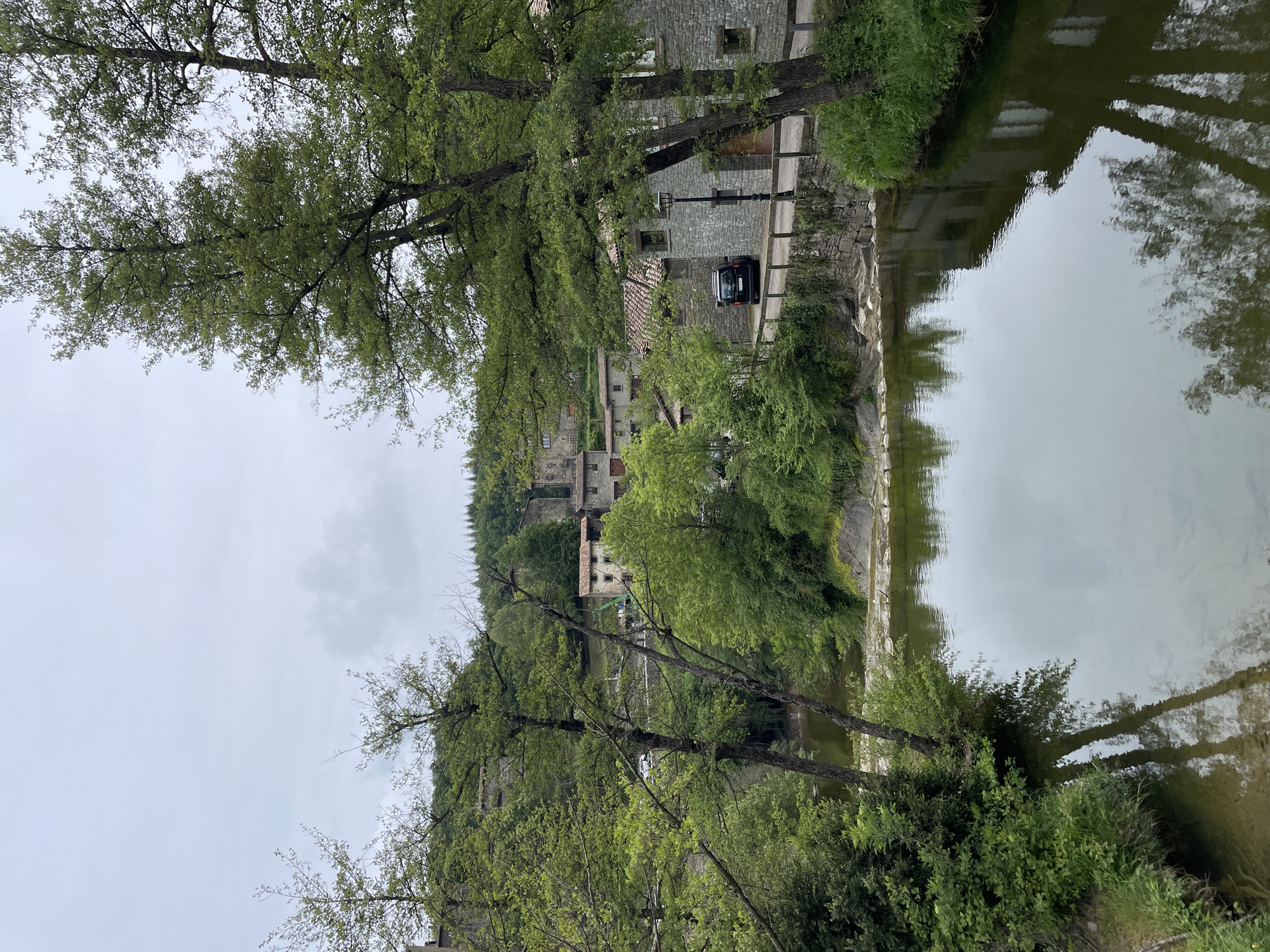 A daytime shot displays a river surrounded by lush vegetation in the municipality of Rupit i Pruit, located in Spain. The river, positioned on the right side of the image, reflects the buildings and trees that border it. Trees line the riverbank, obscuring some of the architecture. Several stone buildings are visible on a hill, some appearing to be residences. A black vehicle is parked next to a building. The sky is overcast, suggesting possible rain or a cloudy day. The image is oriented vertically.