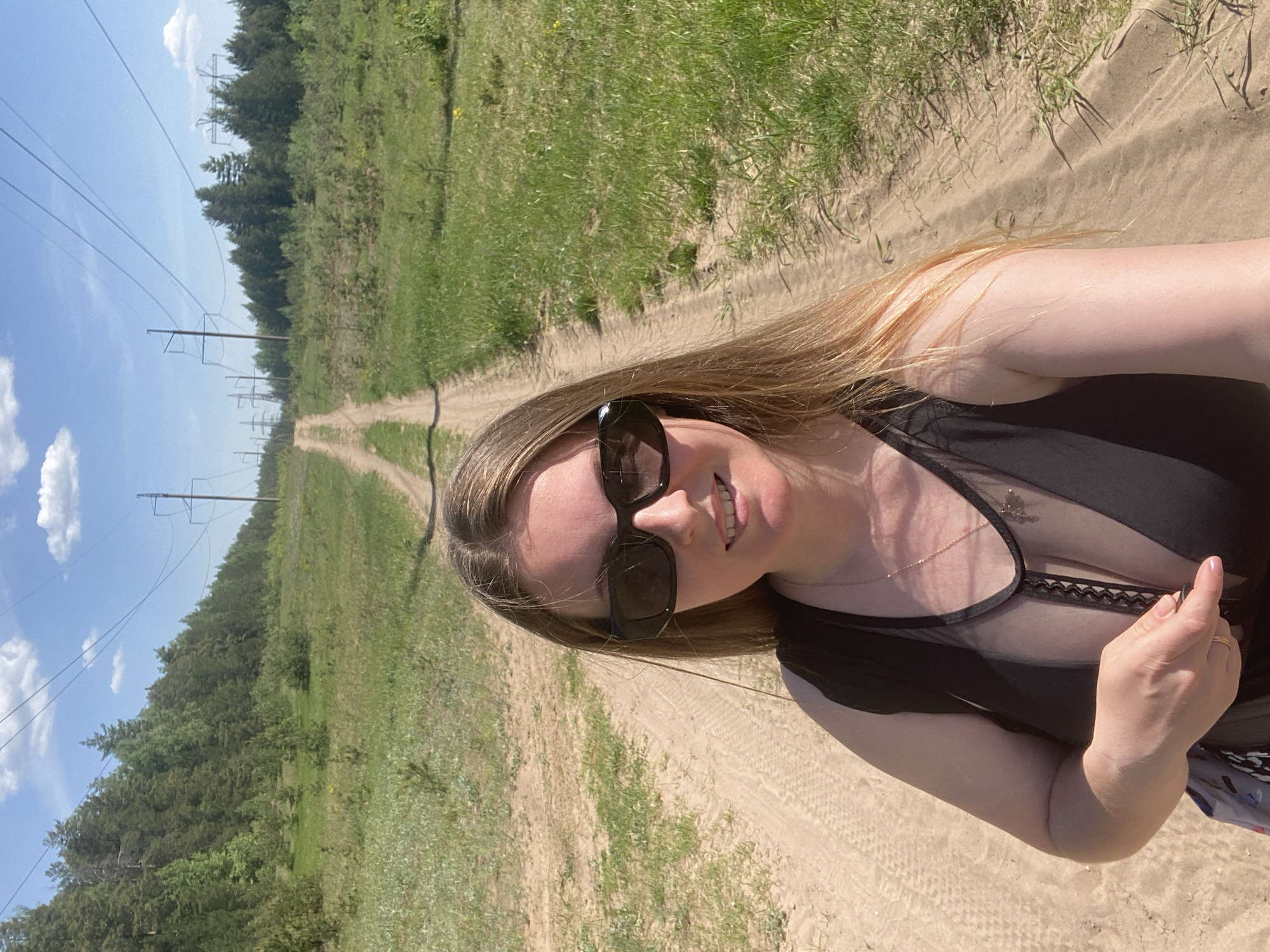 A woman with long blonde hair is outdoors on a sandy dirt road. She is wearing sunglasses and a black top with a sheer neckline. The road stretches into the distance between grassy fields and a line of trees, with power lines running alongside. The sky is blue with scattered clouds. The location is likely Noviy Urengoy, Russia, due to the landscape and the presence of power lines typical of the region. The woman appears to be taking a selfie.