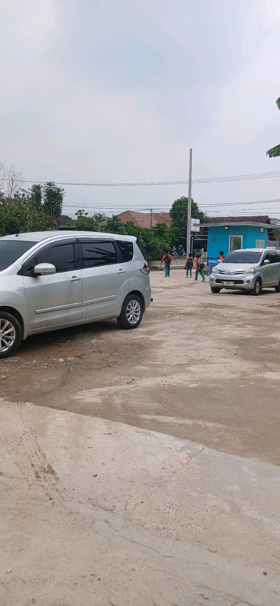 In Metro, Indonesia, a light silver minivan is parked on a dirt and concrete lot. In the background, a smaller silver sedan is driving on the lot. Several individuals are walking in the distance, some carrying backpacks. A small blue building with a white trim and a sign that reads "KEL" is situated behind the individuals. The environment appears to be a somewhat unkempt area with scattered debris and tire tracks on the ground. Trees and houses are visible in the background, and the sky is overcast.