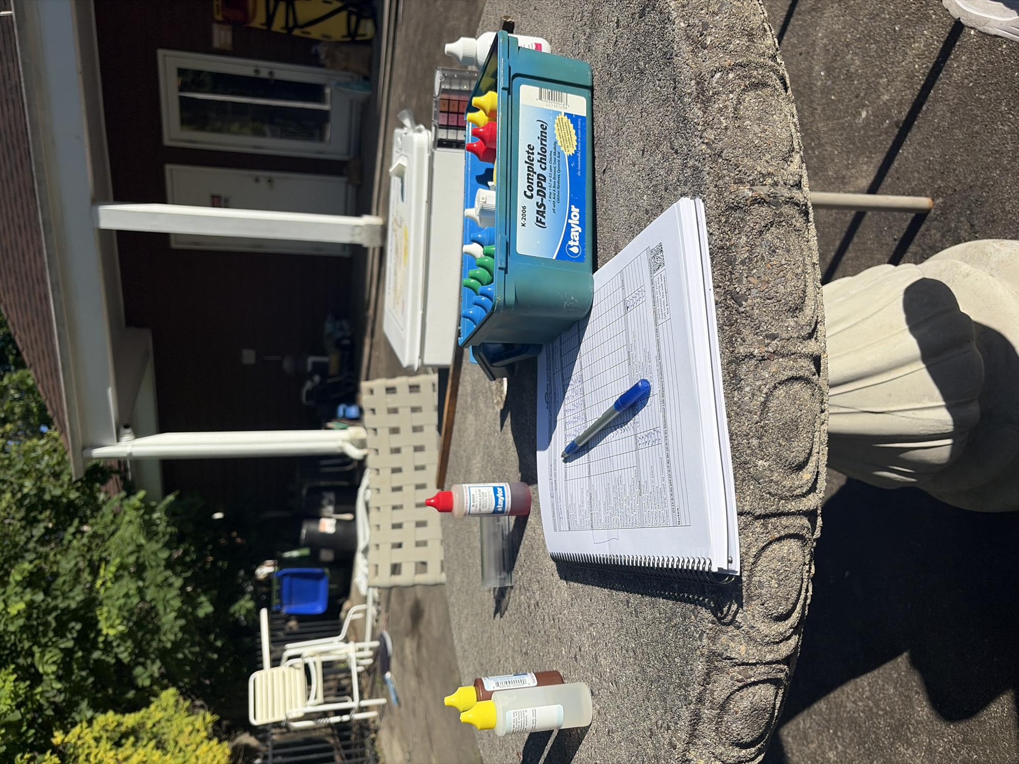 This outdoor scene shows pool maintenance and testing equipment on a textured grey table. The equipment includes a Taylor K-2006 FAS-DPD chlorine test kit in a blue case with various colored caps on reagent bottles, a clear testing vial with a red-capped reagent bottle nearby, and a pair of yellow-capped bottles. A spiral-bound notebook is open next to the equipment, with a blue pen resting on its pages filled with columns and text related to pool testing and maintenance. In the background, there is a building with open windows and white trim. A set of stacked white chairs and a blue plastic tub are visible near a metal fence. Trees and greenery surround the area. The photograph was taken in Edison, United States of America.