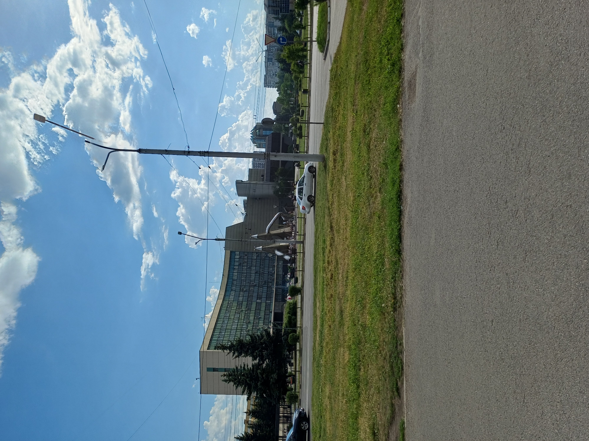 The image shows a daytime scene in a city, featuring a large, curved, multi-story building with many windows. In front of the building, there is a street with cars and a grassy median strip. Trees are planted along the street, and there are streetlights and power lines visible. The sky is blue with scattered white clouds. The image appears to be taken from a street-level perspective, with the foreground dominated by asphalt road.
