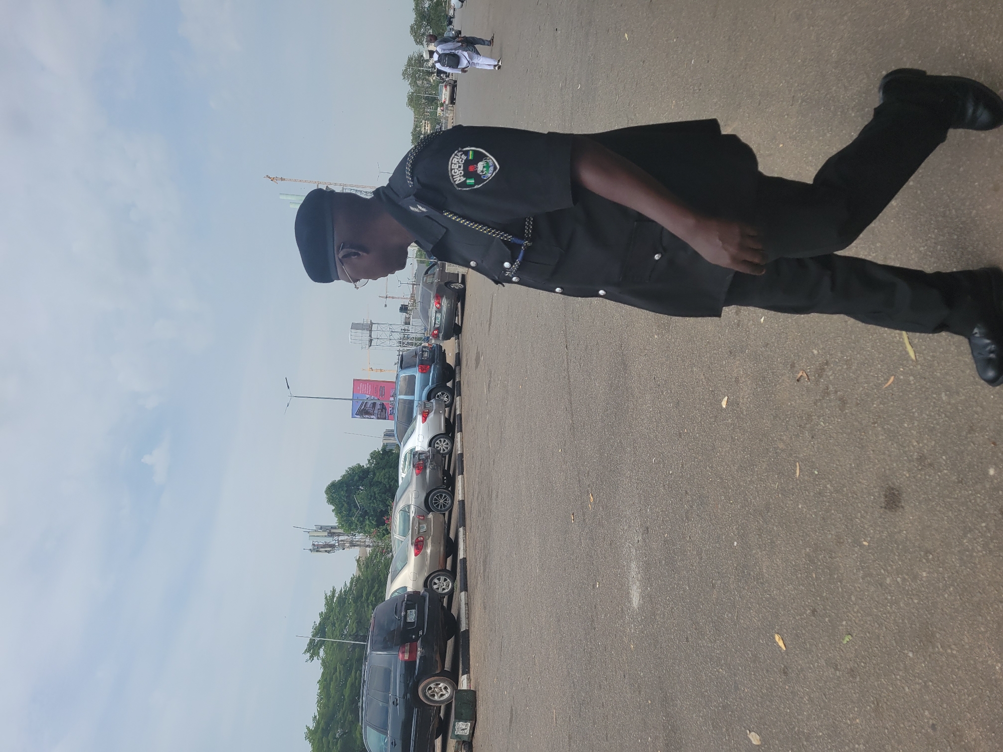 The image shows a Nigerian police officer standing on a road in Abuja, Nigeria. He is wearing a black police uniform with a police patch on his left shoulder. The officer is also wearing a black hat and glasses. Behind the officer are several parked cars. In the background are buildings, trees, and the sky. There are also some people walking in the distance.