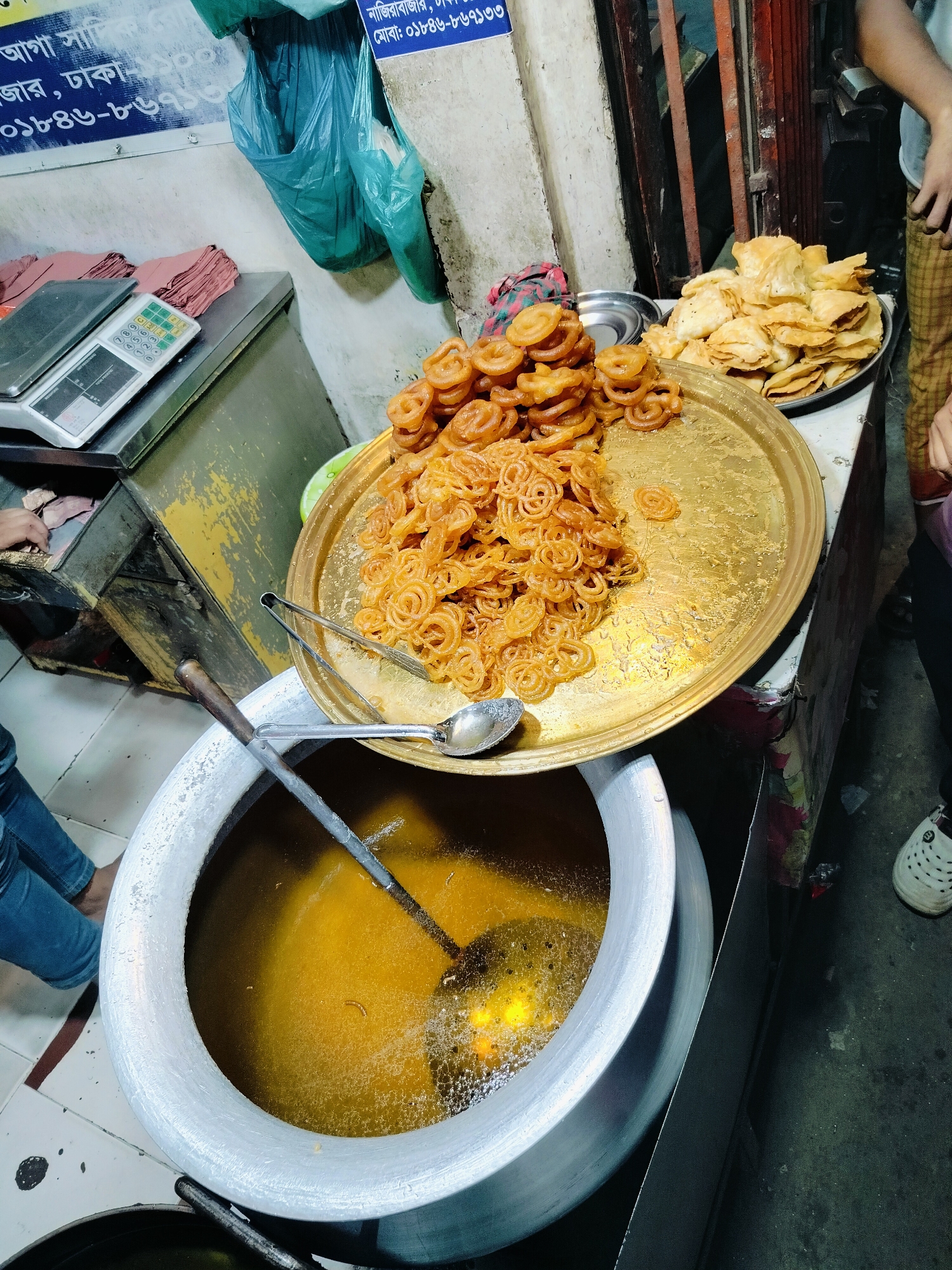 The image shows a street food stall in Dhaka, Bangladesh, likely preparing Jalebi, a popular sweet snack. A large aluminum pot is filled with hot oil, in which Jalebis are being fried. A tray of freshly made Jalebis is placed to the right of the pot, and another tray of what appears to be savory fried pastries sits behind it. A digital scale is visible on a counter to the left, suggesting that the food is sold by weight. A person's legs in blue jeans are visible in the lower left corner, indicating their presence at the stall. Another person's legs wearing checkered pants and a white shoe are seen on the right. The background shows a wall with some signs in Bengali script, and a metal gate. The overall scene depicts a bustling, informal food preparation and vending environment common in Dhaka.