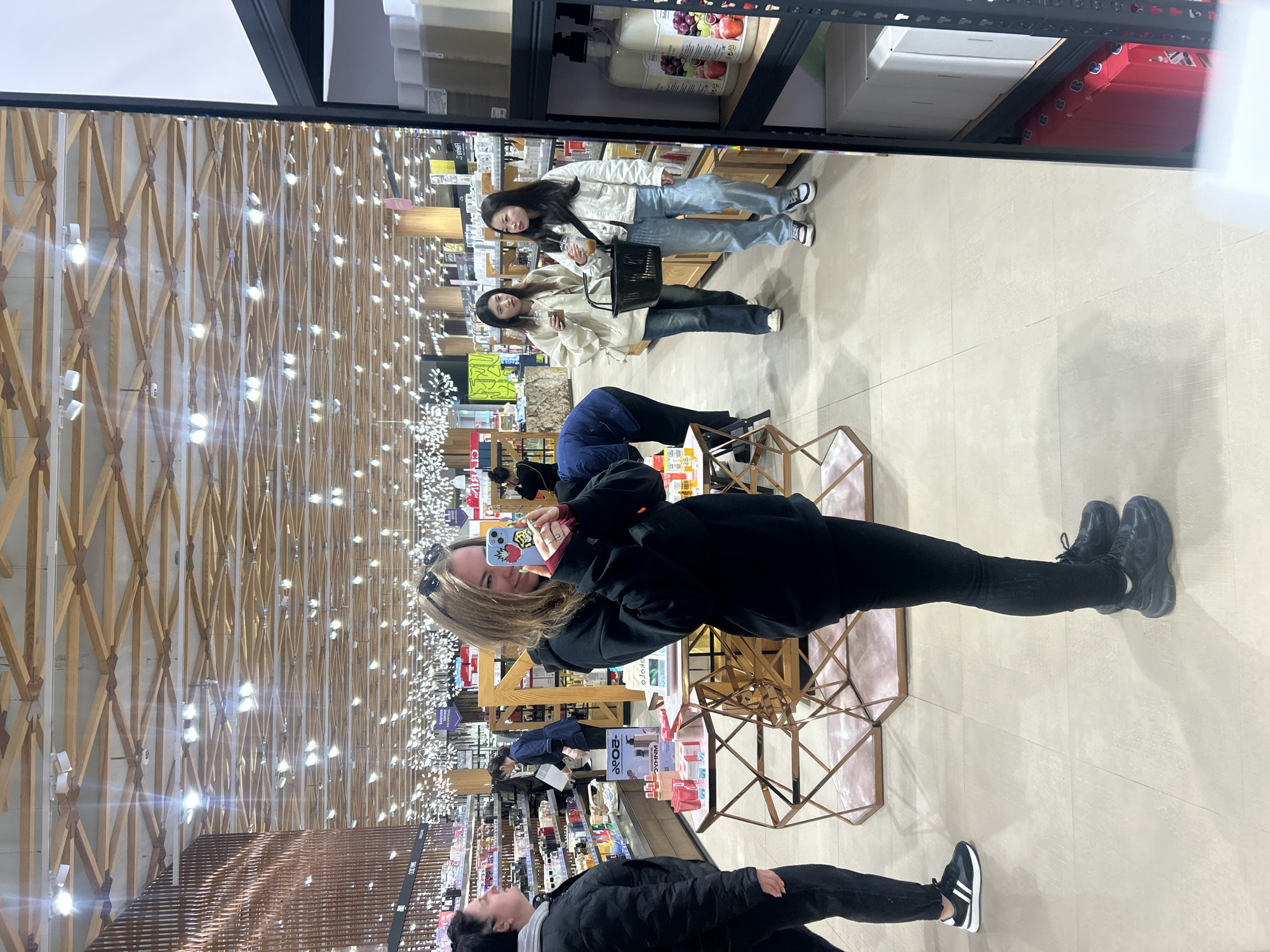 The image shows the interior of a brightly lit retail store. The store's ceiling has a repeating crisscross wooden pattern with small lights attached at each intersection. Aisles are lined with shelves filled with various products.
A person in black leggings, black trainers, and a black hoodie is holding a phone and taking a photo. Reflected in a mirrored surface or glass is a group of three people, two of whom are holding drinks and a black basket. There is another person standing near the bottom of the frame with a gray scarf and a black jacket and trainers. A metallic table or display case is visible near the person taking the photo.
