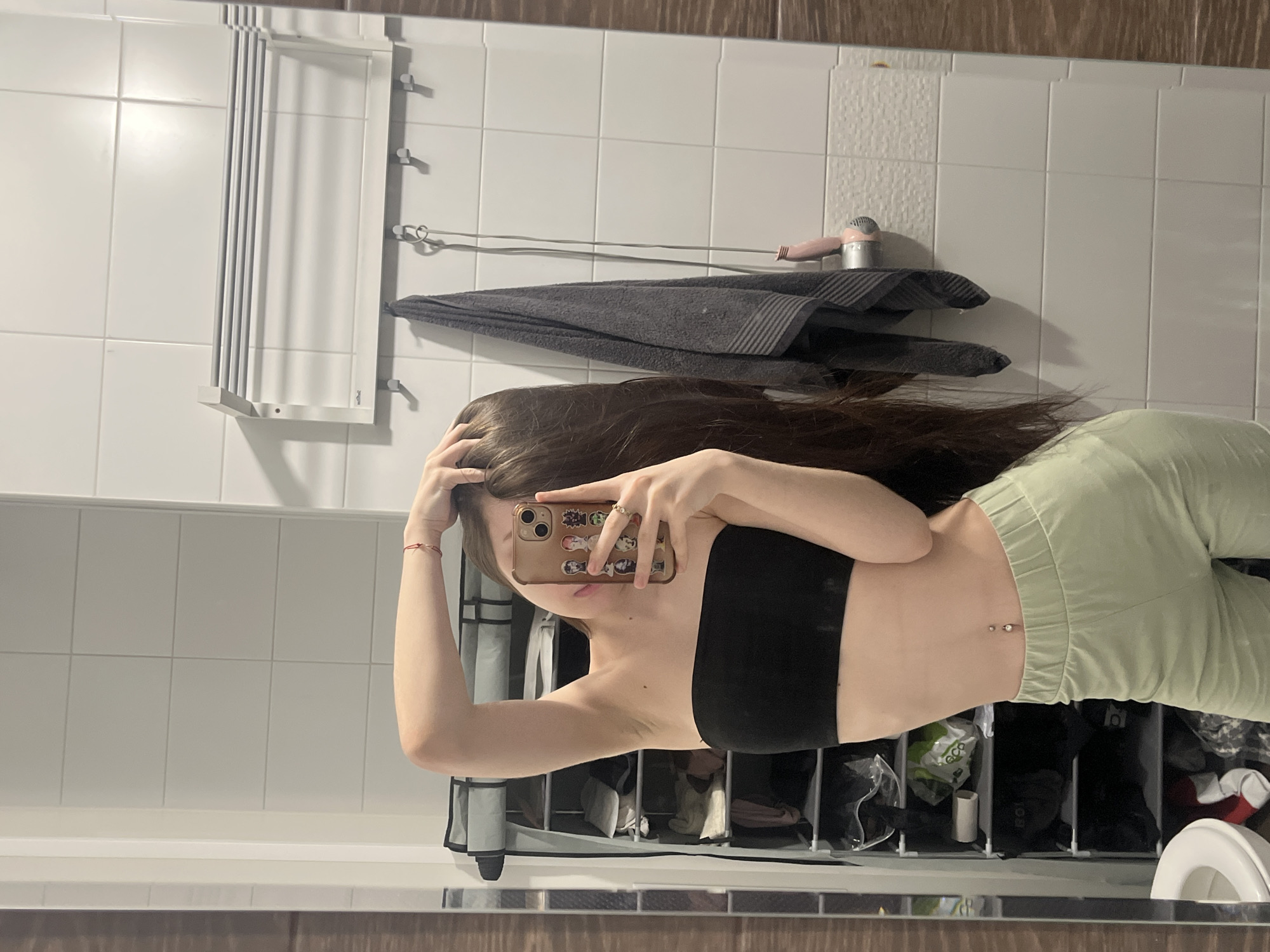 This image is a rotated mirror selfie taken in what appears to be a bathroom in Saint Petersburg, Russia. The person taking the photo, a female with long, dark brown hair, is visible from approximately the chest down to the mid-thigh. She is wearing a black bandeau-style top and light green sweatpants. Her midriff is exposed, revealing a navel piercing. She holds a smartphone with a gold-colored case adorned with multiple anime or cartoon character stickers in her right hand, which also shows rings on her fingers and a red string bracelet on her wrist. Her left hand is partially visible, touching her hair.
The background shows a white tiled wall. To the left, a white wall-mounted folding clothes rack is visible. Above the person's right shoulder, a grey towel hangs on hooks, and a pink and silver hairdryer rests on top of the towel. Below the mirror, a storage unit with multiple compartments contains various items, including bags (one with "ECO" visible text) and other household articles, some appearing to be shoes or accessories. A white toilet bowl is partially visible in the bottom right corner of the reflected scene. The overall lighting suggests an indoor setting, possibly evening, but the exact time of day is not discernible. The image is oriented 90 degrees counter-clockwise from a standard upright view.