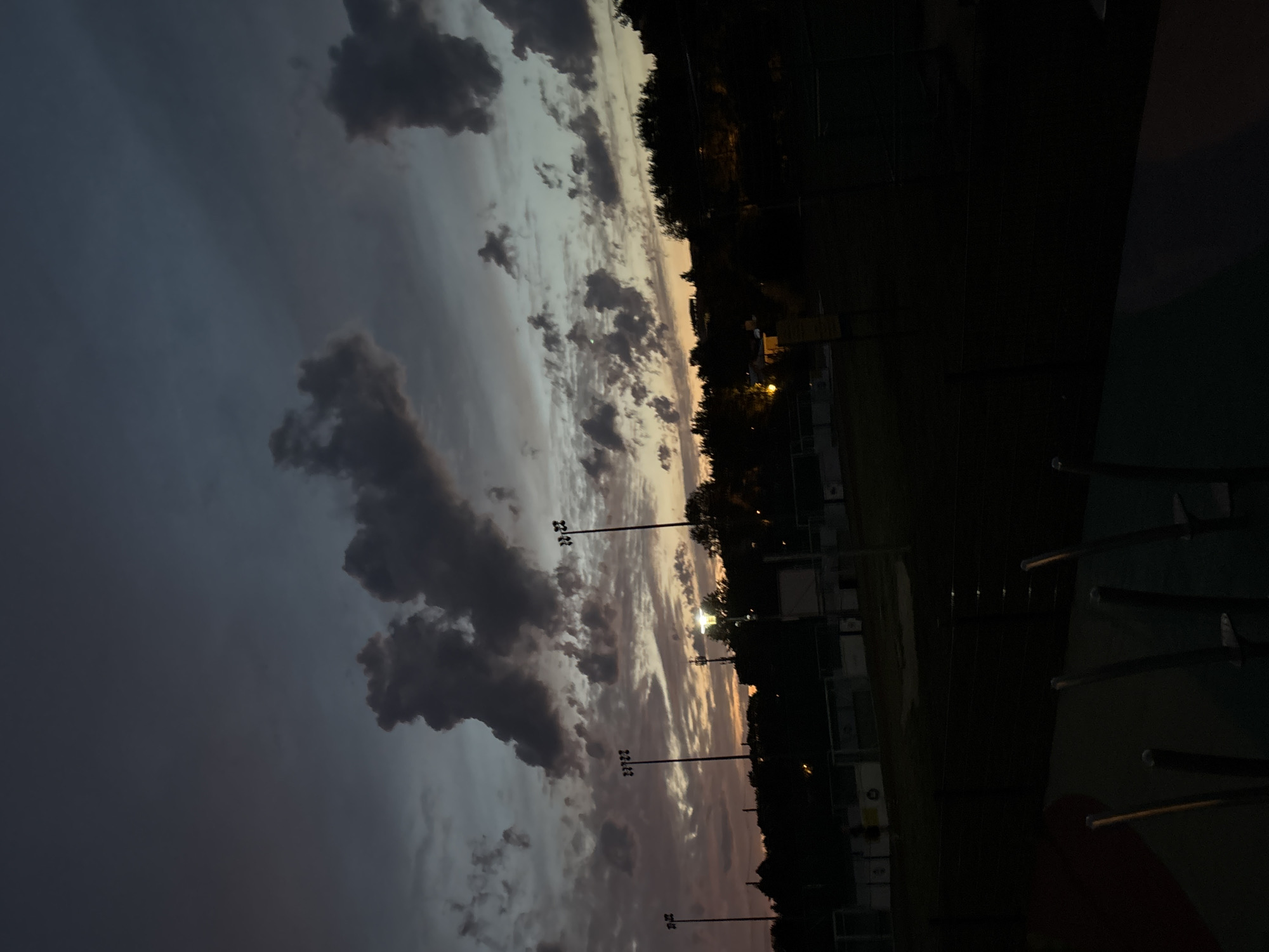 The image shows a dusk sky over a sports field, likely in the city of Lublin, Poland. The sky is filled with dark, textured clouds, with hints of orange and pink near the horizon from the setting sun. Several tall light poles with multiple lamps are visible on the sports field, some illuminated. In the foreground, a dark, out-of-focus area suggests a park or playground with metal structures. The overall scene is dark, with the light poles and the faint glow of the horizon providing the primary sources of illumination. There are no people clearly visible in the image. The landscape appears to be moderately hilly, with trees silhouetted against the sky in the background.