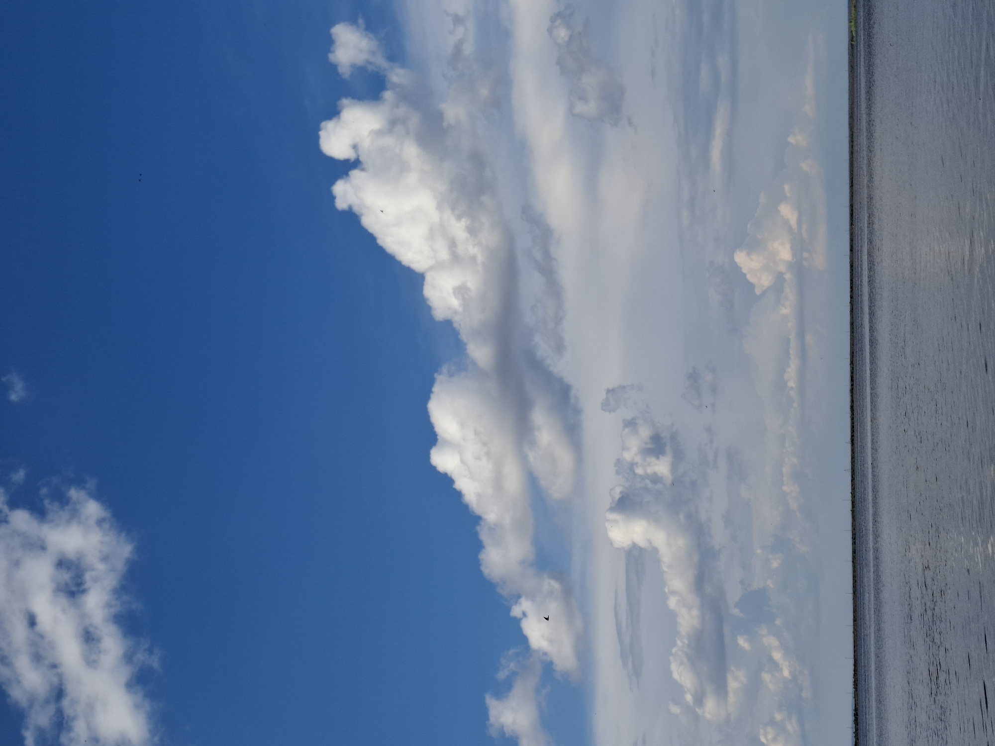 The image depicts a serene landscape with a vast expanse of water under a bright blue sky dotted with white cumulus clouds. The horizon is marked by a faint, distant line of what appears to be structures, possibly wind turbines or utility poles, suggesting a man-made presence against the natural elements. The water's surface is calm, reflecting the sky and clouds, with subtle ripples indicating a gentle breeze. The overall impression is one of tranquility and open space. This scene is likely located in or near the city of Chaikovsky, Russia, a region known for its natural beauty and proximity to large bodies of water, such as the Kama River reservoir.