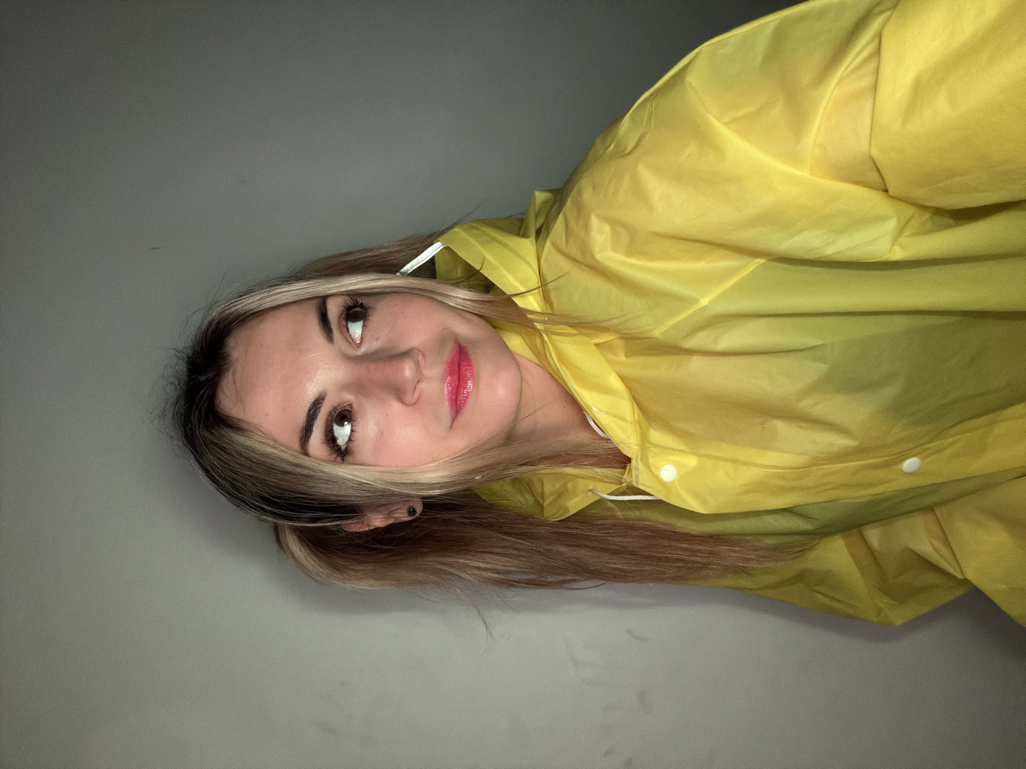 The image is a close-up, chest-up portrait of a young woman, shown rotated 90 degrees counter-clockwise from what would be a natural upright viewing angle. She has long, straight or slightly wavy blonde and light brown hair with prominent lighter highlights framing her face. Her brown eyes look upwards and slightly to her right, displaying a gentle, perhaps contemplative expression. Her lips are slightly parted, adorned with vibrant pink or red lipstick. Her eyebrows are well-defined, and a small black stud earring is visible in her left ear.
She is dressed in a bright, vivid yellow raincoat or jacket, made from a lightweight, somewhat crinkly, waterproof material. White snap buttons are visible on the front, along with a white drawstring or cord near the neckline. The fabric of the raincoat creates soft folds and creases, particularly prominent on the right side of the frame.
The background is a uniform, plain light grey wall or surface, offering no specific details. The lighting is soft and diffused, illuminating her face and the raincoat without harsh shadows, suggesting an indoor environment or controlled lighting conditions. There are no visible text elements, specific location identifiers, or weather cues within the image itself. The overall mood is calm and serene.