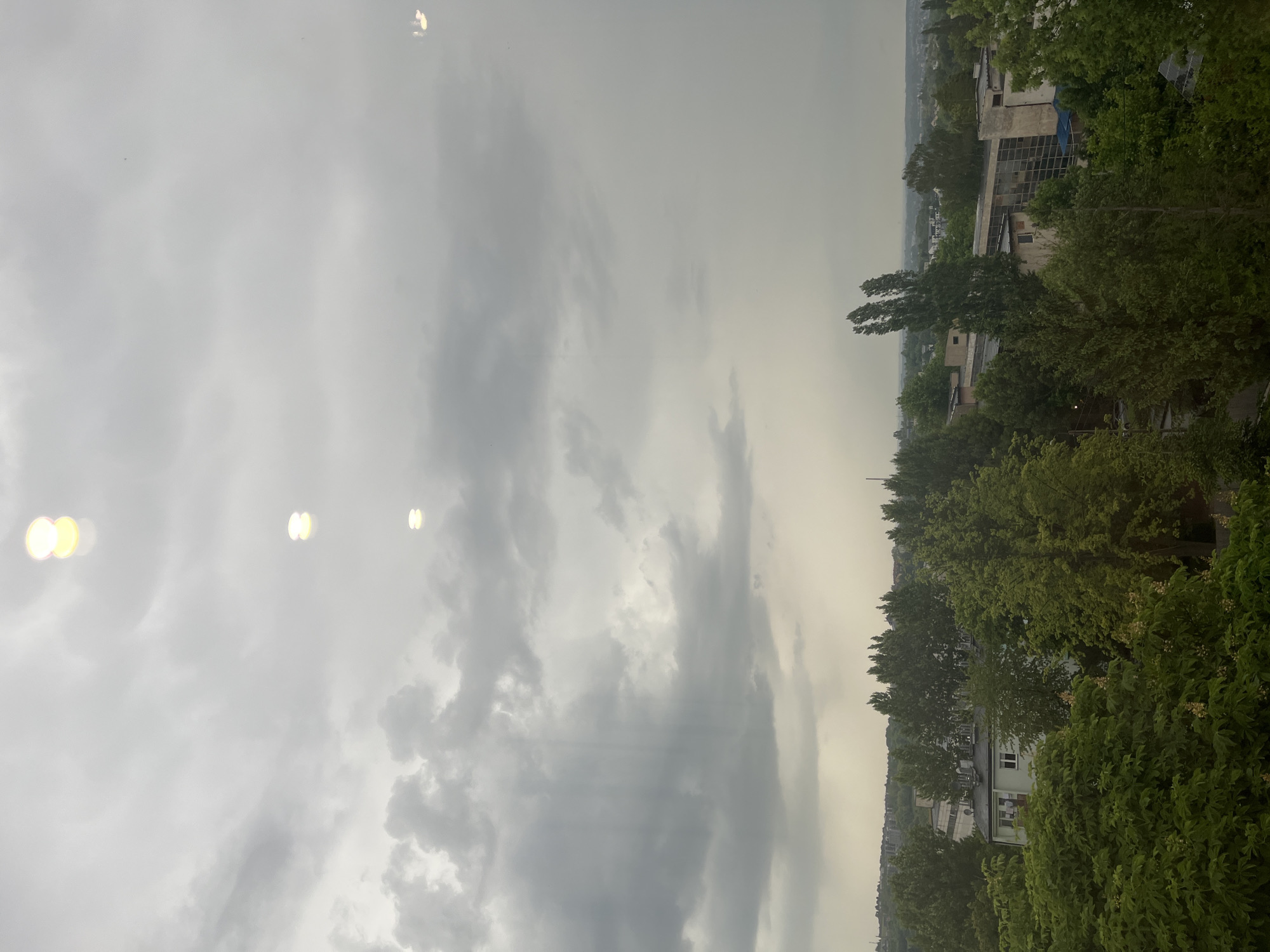 The image depicts an overcast cityscape. The sky is filled with gray clouds and a few bright spots. In the foreground, there is lush green foliage that obscures buildings. Further back, rooftops and building structures are visible, indicating a densely populated area. The location appears to be a city, with an uncertain state and location.
