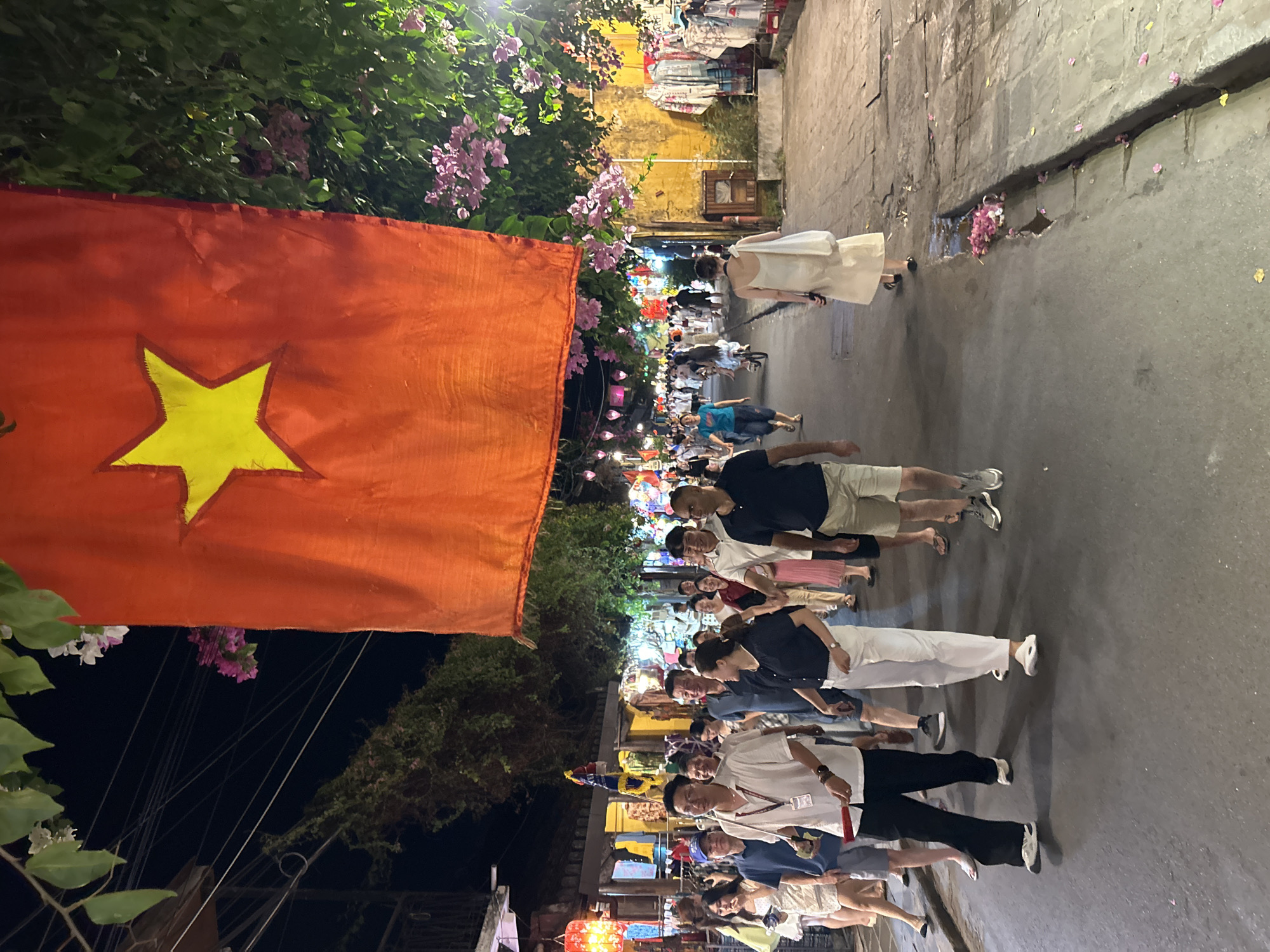 In Hội An Ward, Vietnam, a street is illuminated at night by ambient lighting and hanging lanterns. A large red Vietnamese flag with a yellow star is prominently displayed, draped from above. A crowd of people, appearing to be tourists and locals, walk along the paved street. Some are engaged in conversation, while others are observing their surroundings or walking individually. Shops with goods displayed on racks line the right side of the street. The ground is scattered with fallen pink flowers.