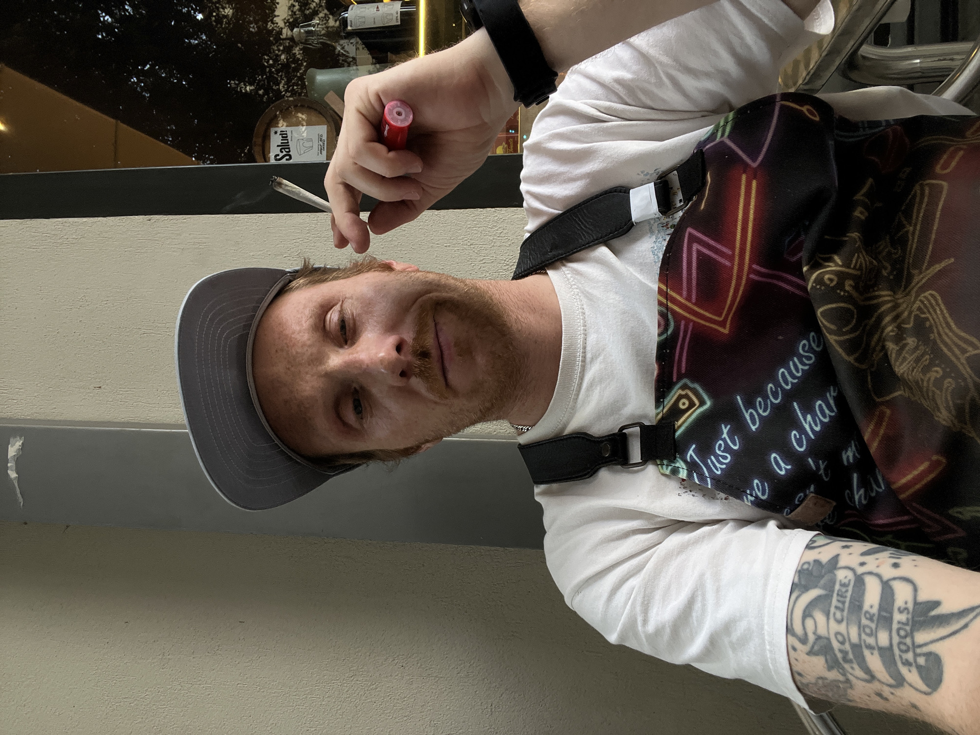 A man with freckles and a gray baseball cap is holding a lit cigarette in his left hand and a red lighter in his right. He is wearing a white long-sleeved shirt and a black apron with colorful patterns. The apron has writing that includes the phrase "Just because we are a char..." on it. He has a tattoo on his left forearm with the words "NO CURE FOR FOOLS." A black smart watch is visible on his left wrist.
In the background, a window reflects outdoor scenery and displays bottles. A "Salut!" sign is visible behind the man's head. Based on the "Salut!" sign and the overall urban atmosphere, the location is likely a bar or restaurant in Tel-Aviv, Israel.