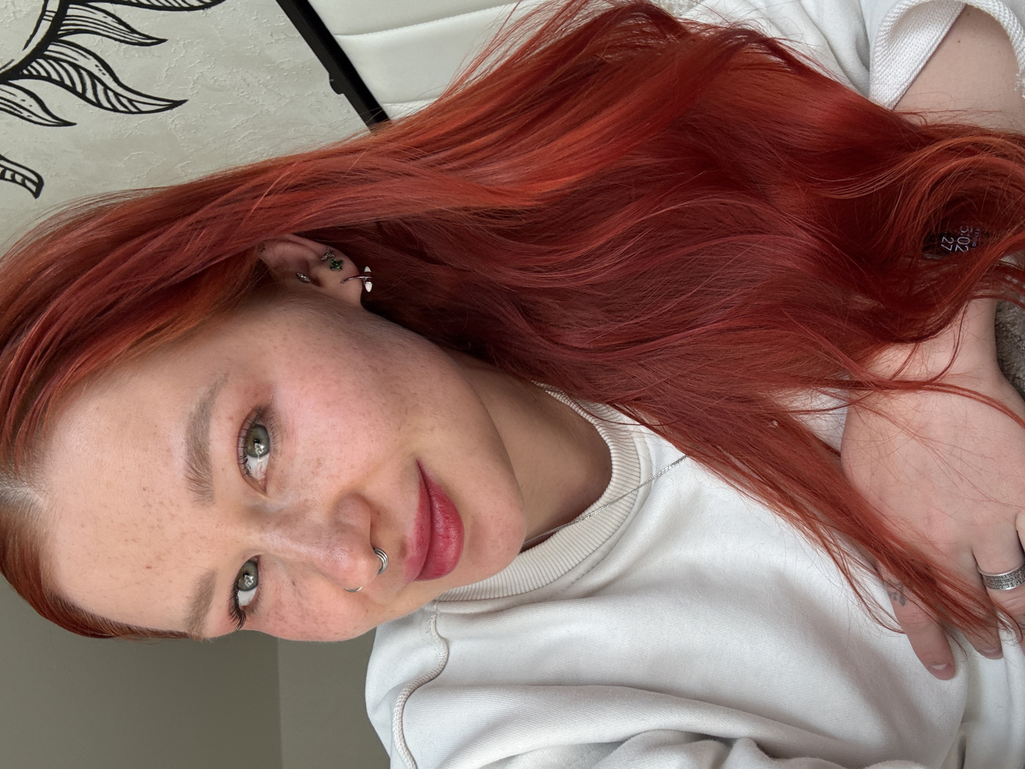 The media features a close-up portrait of a young woman with vibrant red, long hair, fair skin, and numerous freckles, taken in what appears to be an indoor setting in Rostov-on-Don, Russia. She has striking green eyes with subtle eye makeup and lightly colored eyebrows. Her lips are painted a soft red/pink shade, and she has multiple facial piercings, including a double silver hoop in her septum and several earrings in her left ear, one of which is a small green clover design.
She is wearing a casual white crewneck sweatshirt and a thin silver chain necklace. Her right hand, adorned with a silver ring displaying partially visible text, rests near her body. Her expression is calm with a gentle, soft smile as she looks directly towards the camera. The background reveals a decorative, possibly textile, item with a black and white leaf-like or sunray pattern. A small black tag with white text "S.02" and "27" is visible within her hair on the right side of the image.