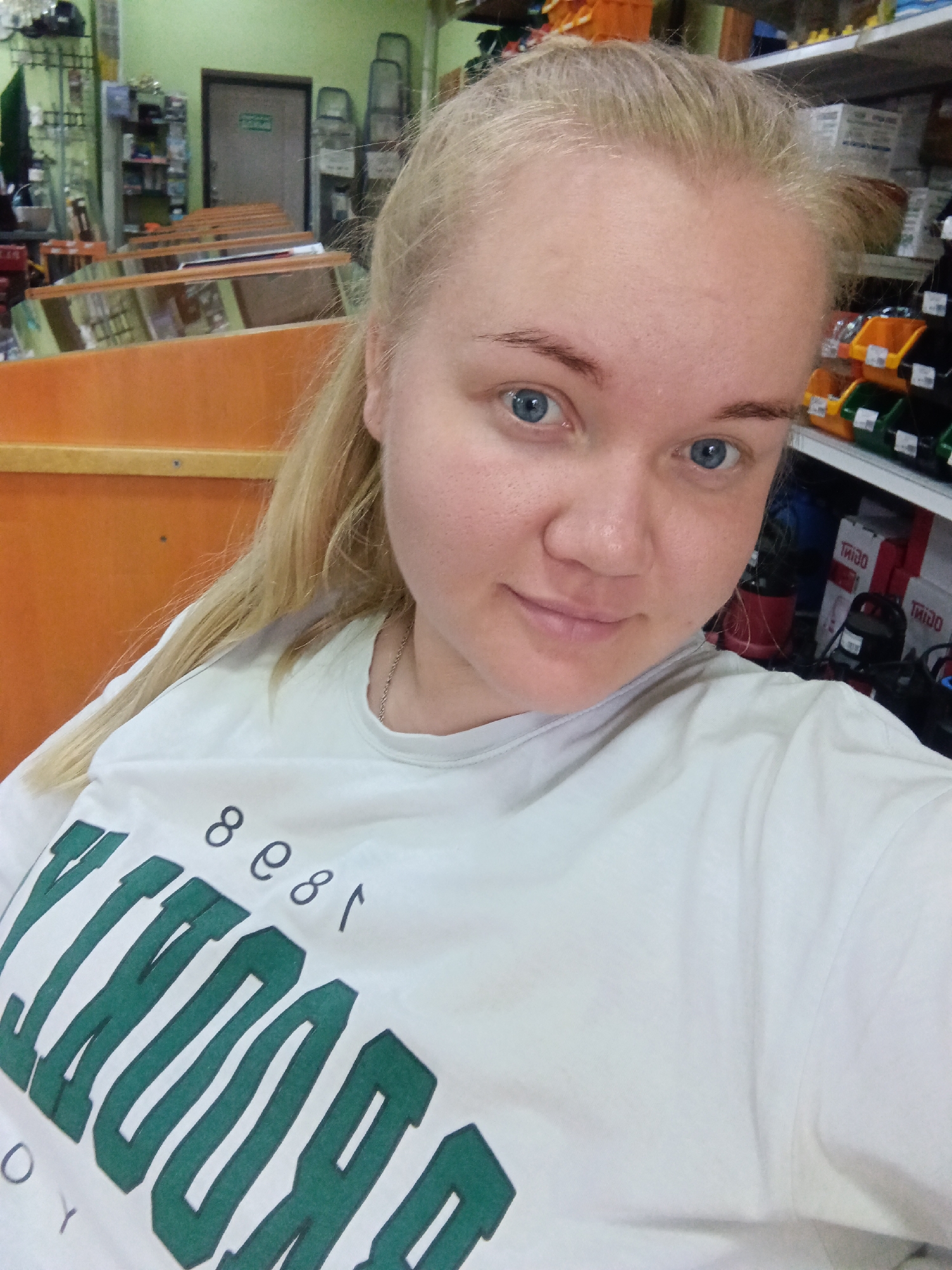 In the city of Фоки, Russia, a young blonde woman with blue eyes and fair skin is taking a selfie in what appears to be a retail or storage environment. She is wearing a white t-shirt with "BROOKLYN" printed in large green letters and the numbers "8 81" in black above it. Behind her, shelves are visible, stocked with various items including what appear to be boxes and bins. To her left, there are orange-colored display units or shelves, and further back, a door with a green sign is visible. The lighting suggests an indoor setting, possibly a warehouse or a store.