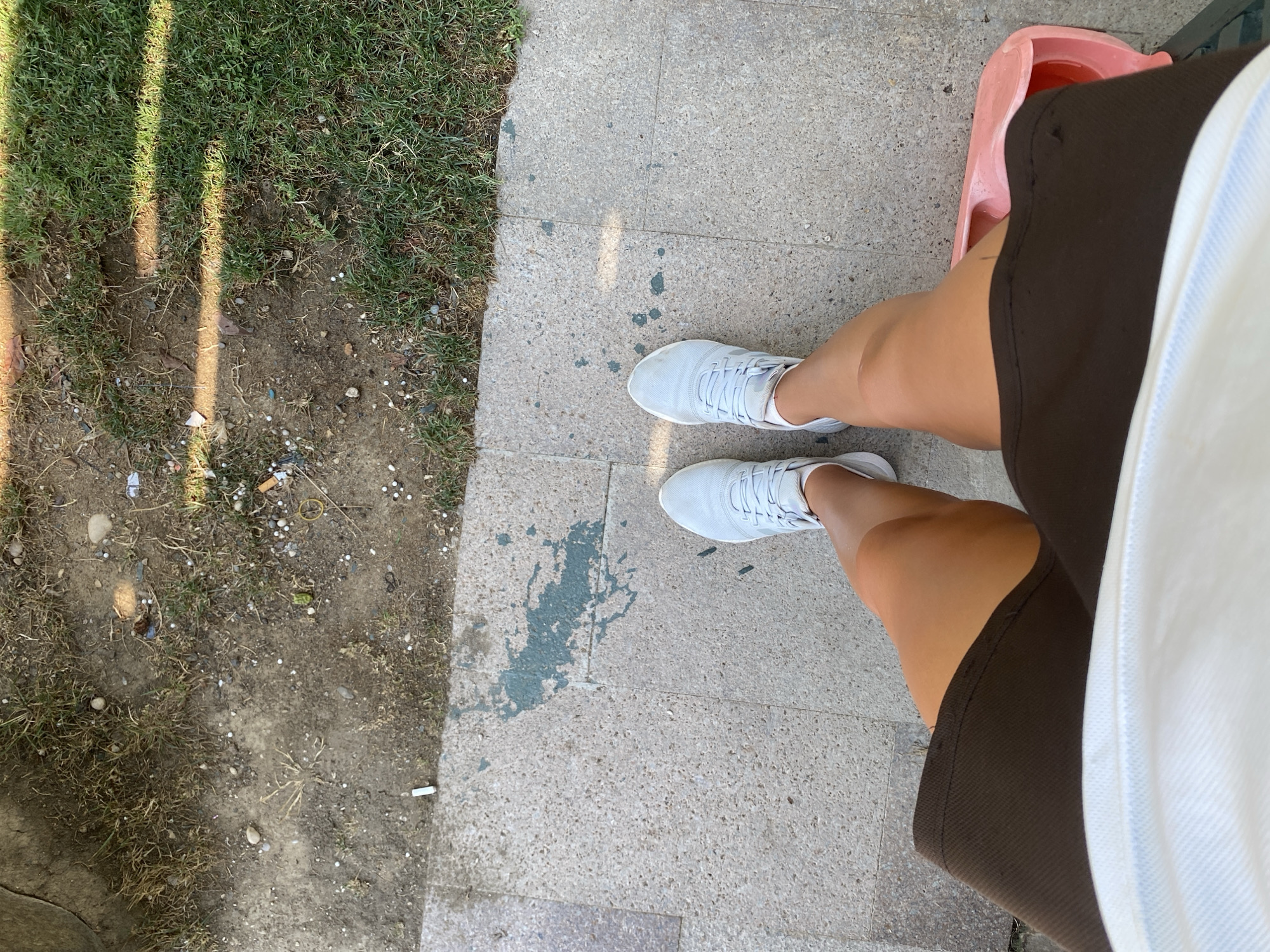 This is a high-angle, full-body shot of a person's legs from the knees down. The person is standing on a paved area with scattered patches of grass and dirt. They are wearing white athletic shoes and brown shorts. In the background, there are glimpses of grass and dirt with some debris, possibly cigarette butts. The paved surface has some greenish stains. To the right, a pink object, possibly a plastic container, is partially visible. The lighting suggests it is daytime, with shadows falling across the scene. The location is identified as Aksu, Turkey, though no specific landmarks are visible in the image.