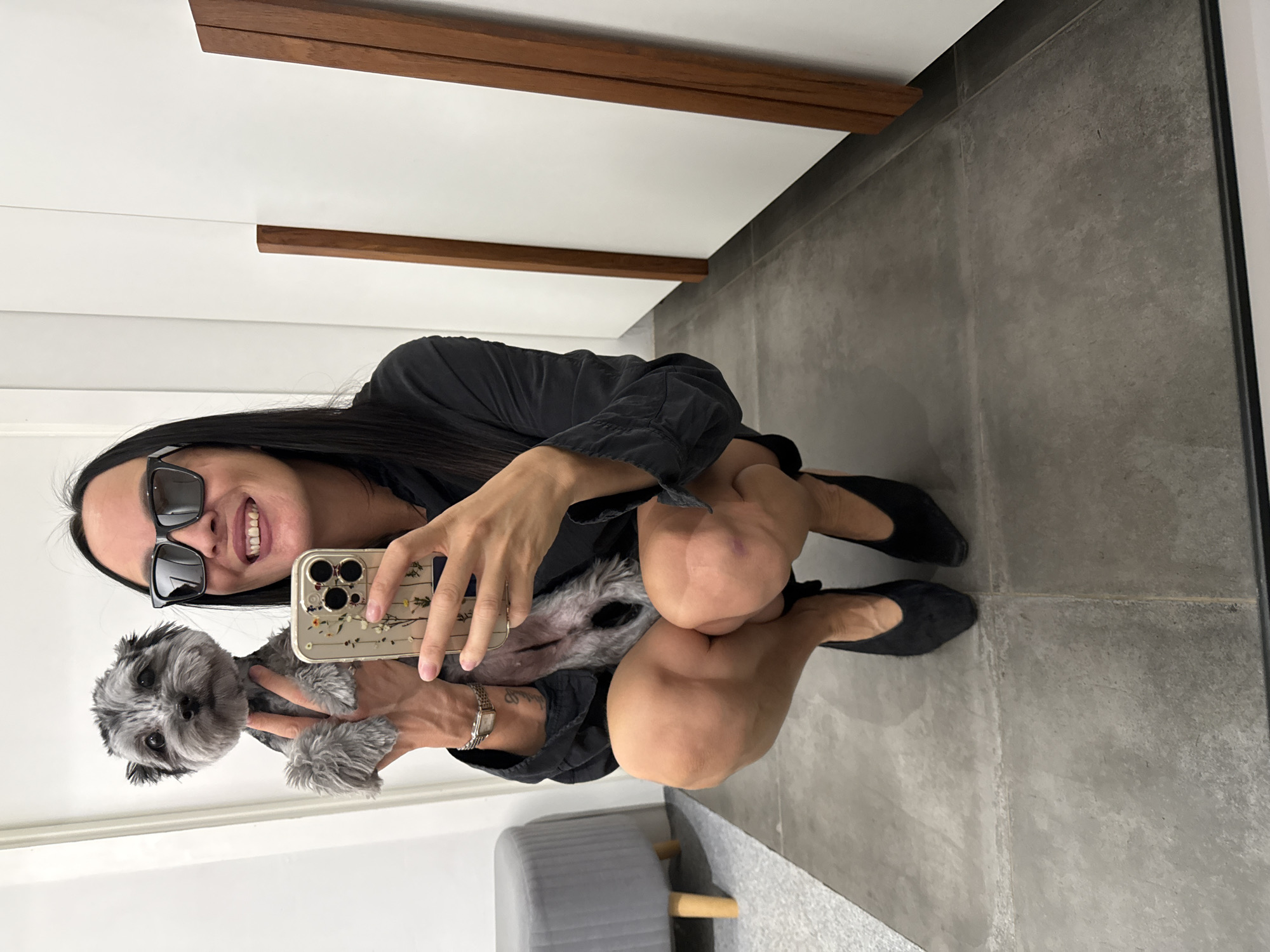 A woman with long, dark hair and black sunglasses is squatting down, holding a small, grey dog. She is taking a selfie with her phone, which has a floral case. She is wearing a black shirt and black heels. The background appears to be a dressing room or fitting area, with white walls, wooden accents, and grey tiled flooring. There is a grey ottoman visible in the background to the left. The image is devoid of any specific geographical markers that would associate it with Chaikovsky, Russia, or any other city.