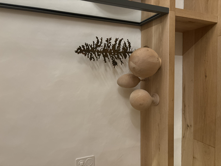 The image shows a wooden shelf with three wooden spheres and a metal silhouette of a pine tree on top. The shelf is a light wood color and is mounted on a wall. The wall is painted a light beige color and has a white electrical outlet on the left side. The shelf is part of a larger wooden structure. The wooden spheres are all different sizes and shapes, and the silhouette of the pine tree is in the center of the shelf. The image is taken from a low angle, looking up at the shelf. It is located in a building in the city of Celerina/Schlarigna, Switzerland, at coordinates 46.514, 9.859.