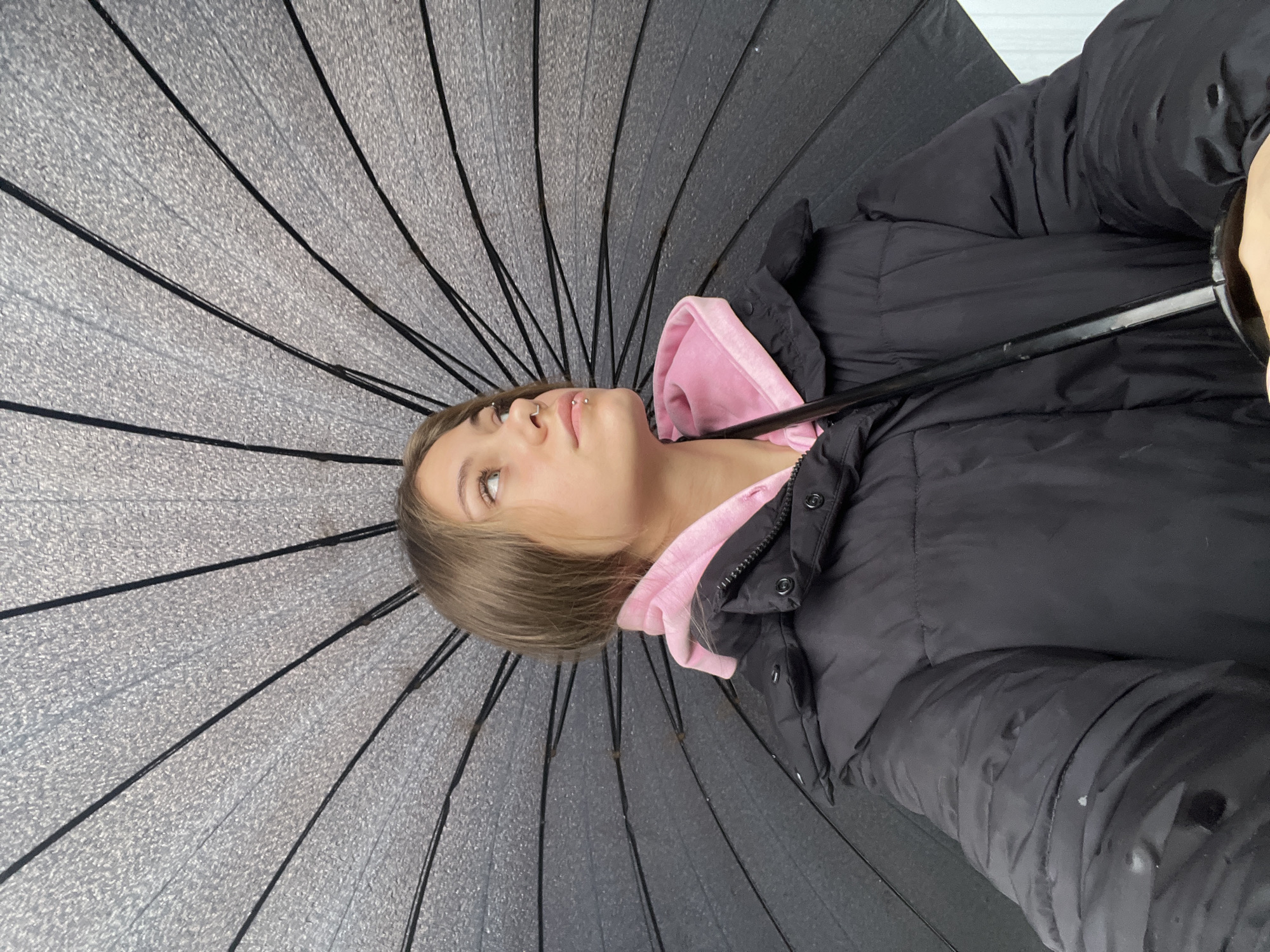 A young woman stands under a black umbrella. She is wearing a black jacket with a pink hoodie underneath. She has fair skin, light brown hair, and piercings on her nose and lip. The umbrella has a textured gray canopy and black ribs. The background is out of focus, but appears to be a building or wall. The location information is Фоки, Russia. The image has been rotated counter-clockwise.